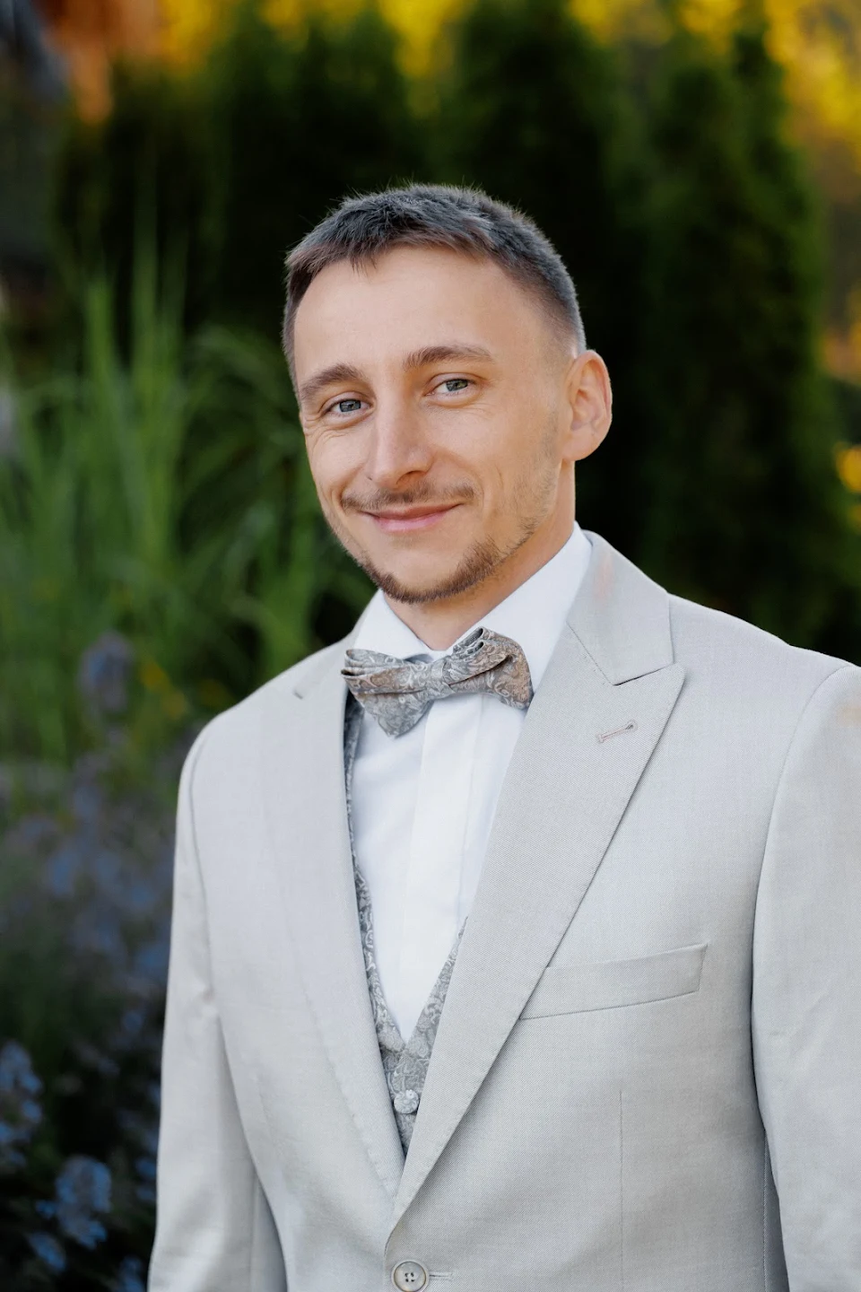 Smiling man wearing a light gray suit with a white shirt and patterned bow tie, standing outdoors with greenery behind him.