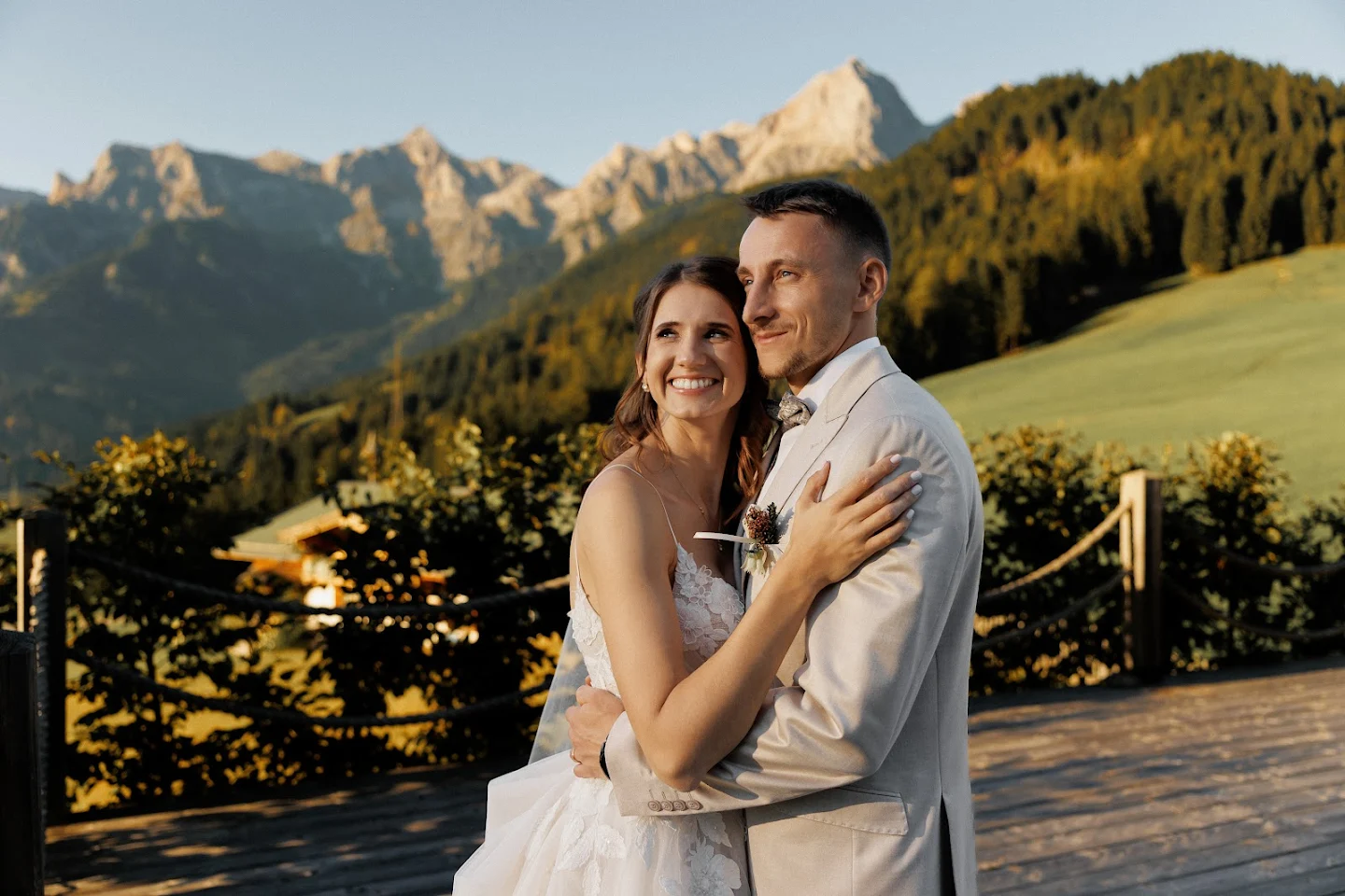 Bride and groom embracing outdoors with mountains and forest in the background during golden hour.