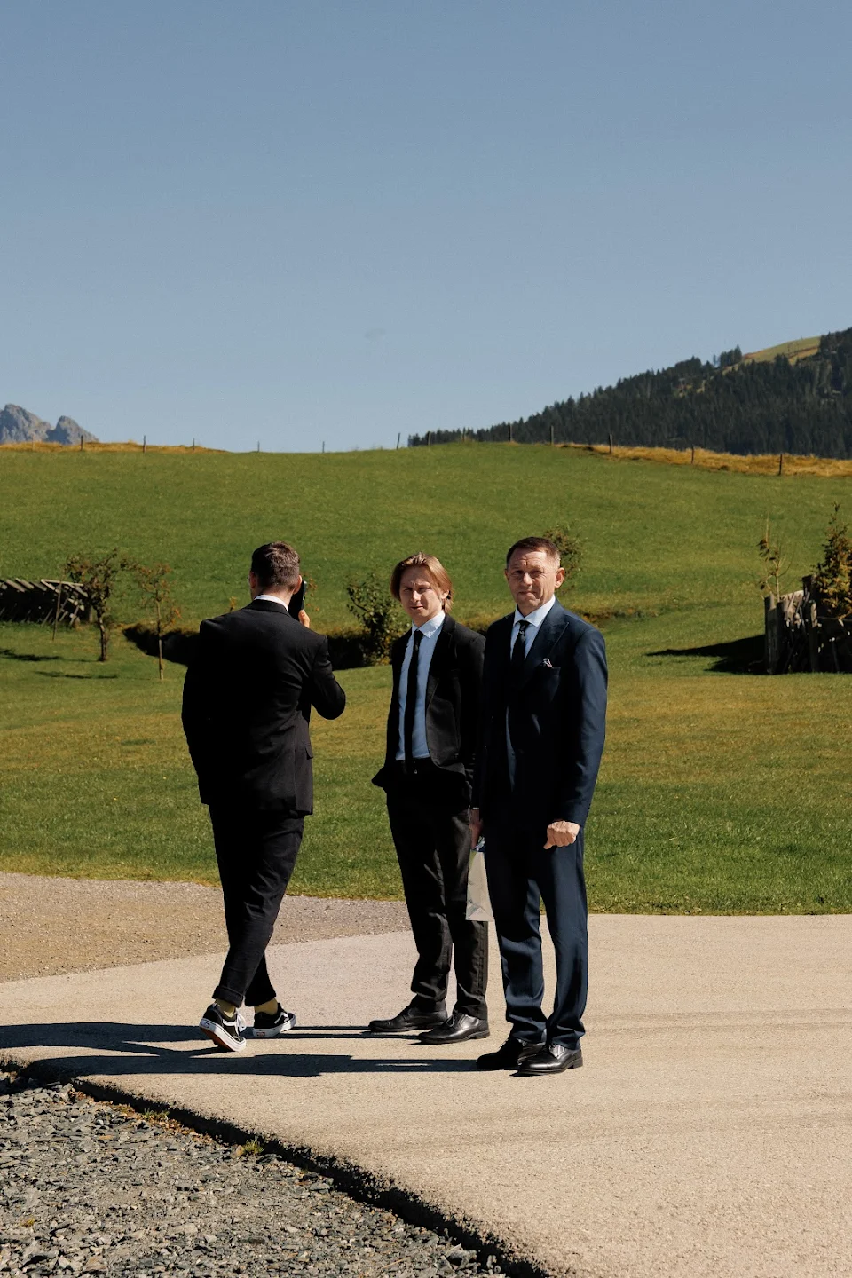 Three men in suits standing on a paved path by green hills under a clear blue sky.