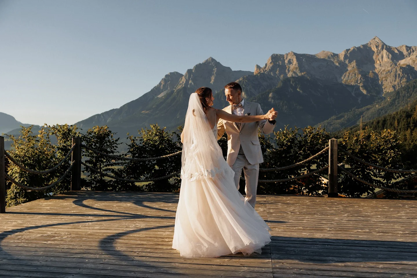 Bride and groom dancing on a wooden deck with mountain landscape in the background during sunset.