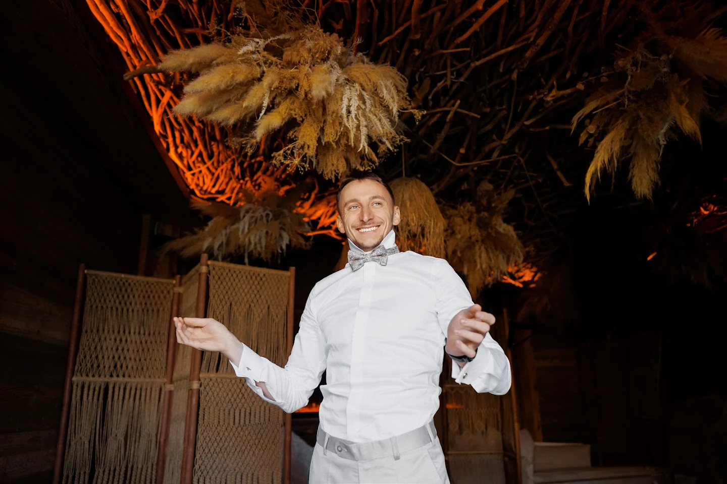 Smiling man in white shirt and gray bow tie standing indoors with decorative dried plants and woven partitions in warm lighting.