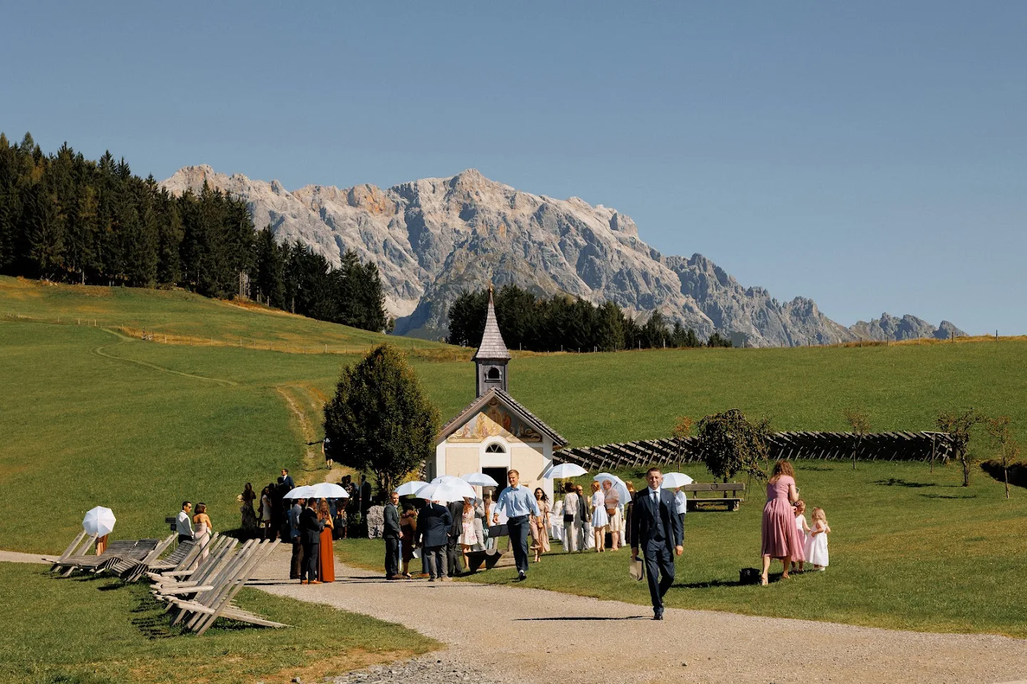 People gathered outside a small chapel in a green mountainous landscape with some holding white umbrellas.