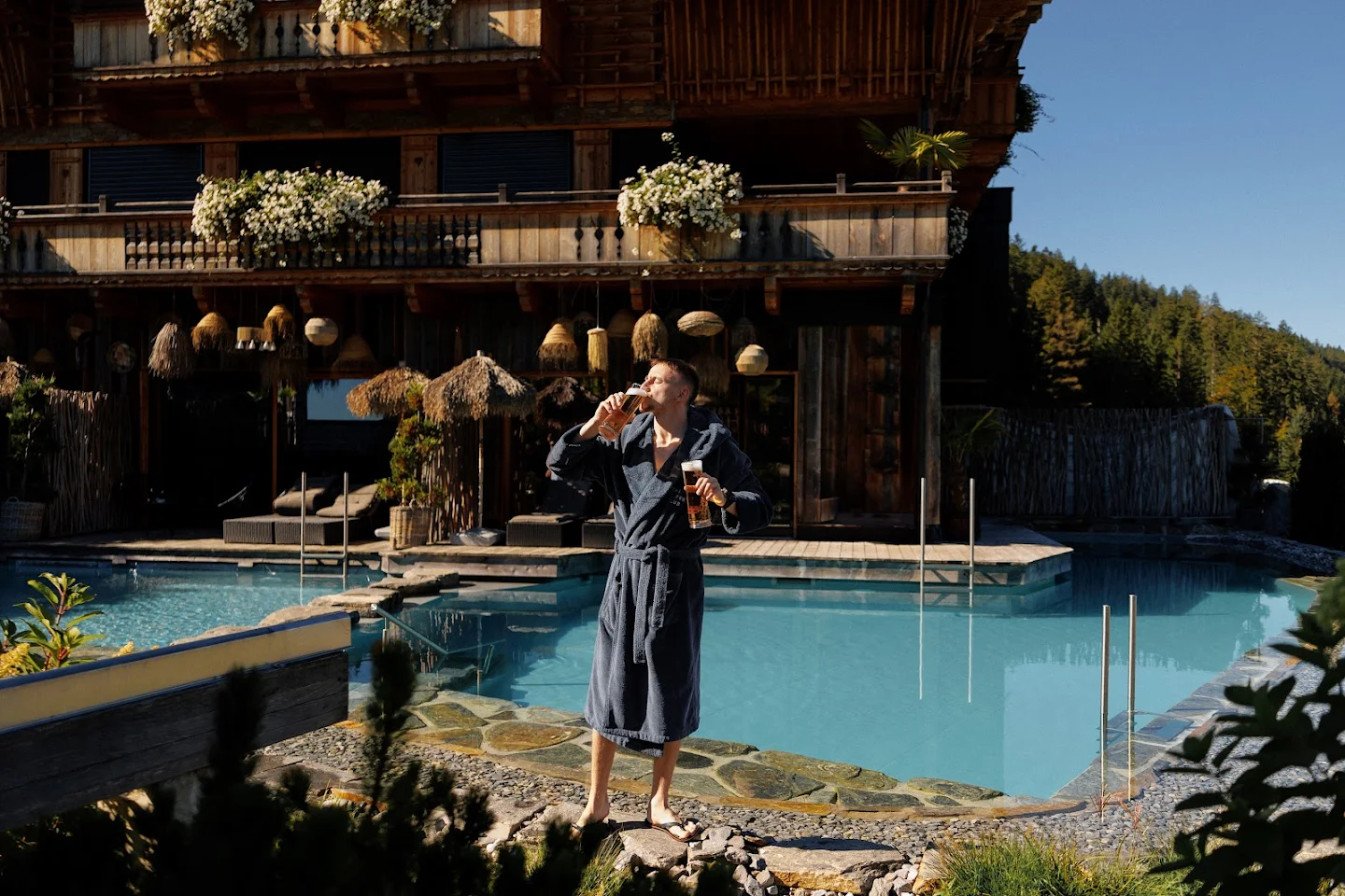 Man in a dark bathrobe standing by a pool drinking beer with a rustic wooden building and greenery in the background.