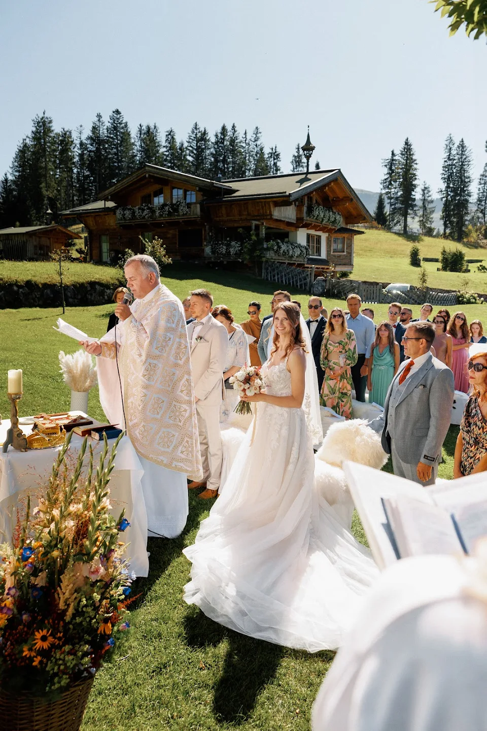 Bride and groom standing outdoors at a wedding ceremony with a priest speaking into a microphone and guests watching.