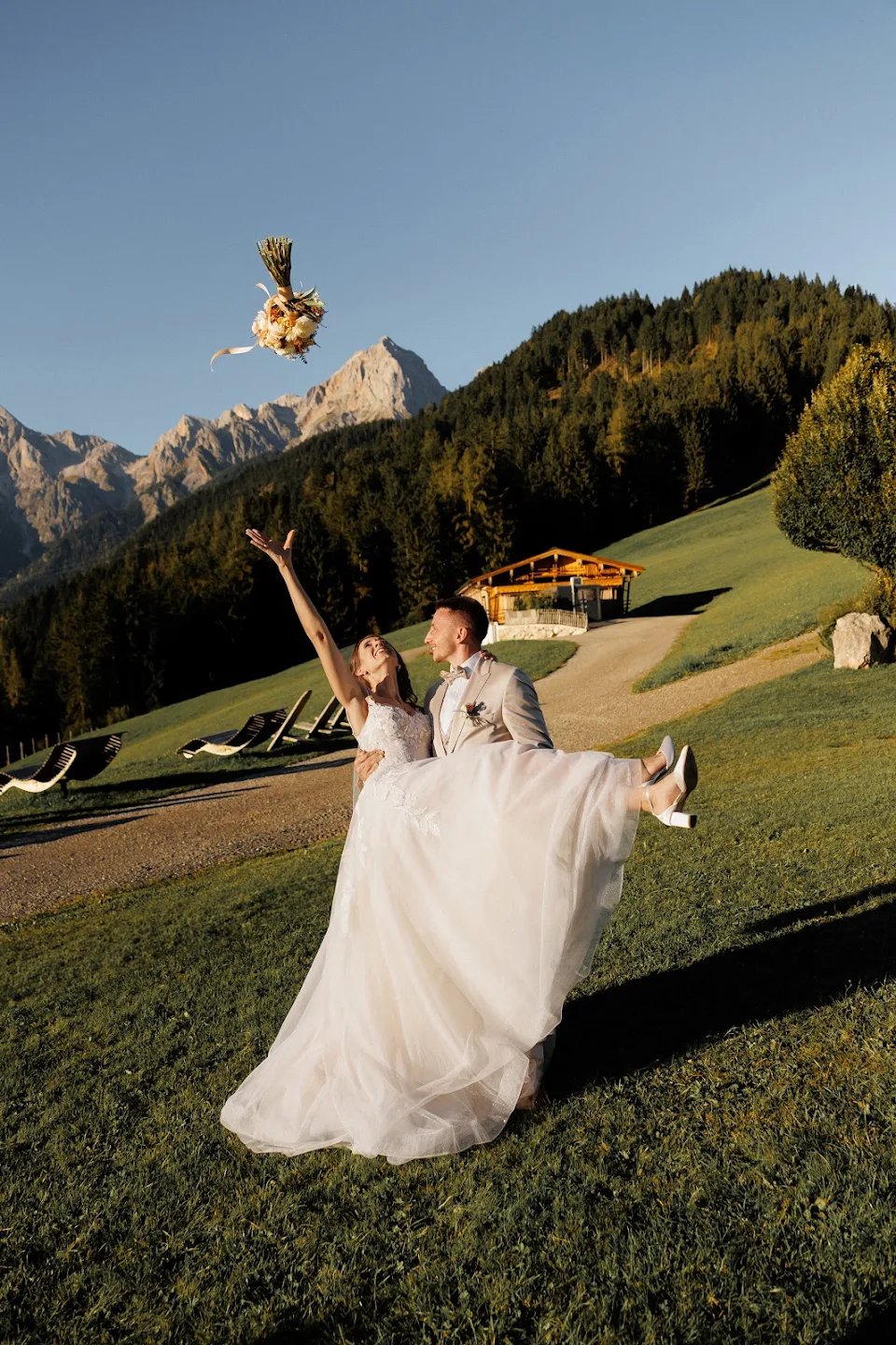 Bride in a white dress being carried by the groom outdoors, tossing a bouquet in the air with mountains and trees in the background.