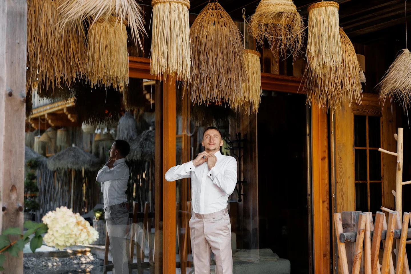 Man buttoning the collar of his white shirt standing in front of a glass door with woven straw lamps hanging above.