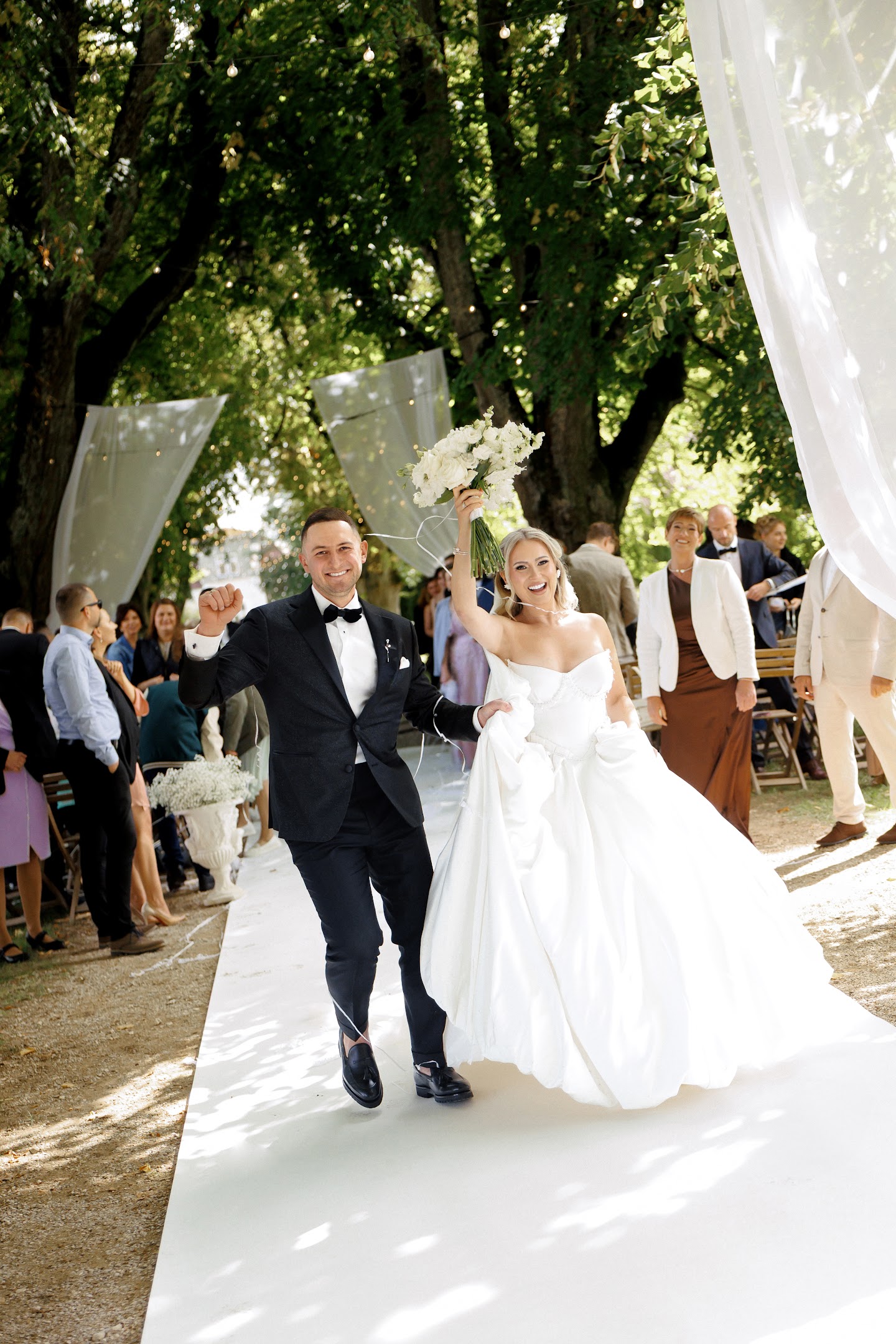 Bride and groom joyfully walking down outdoor aisle with bride holding bouquet up high.