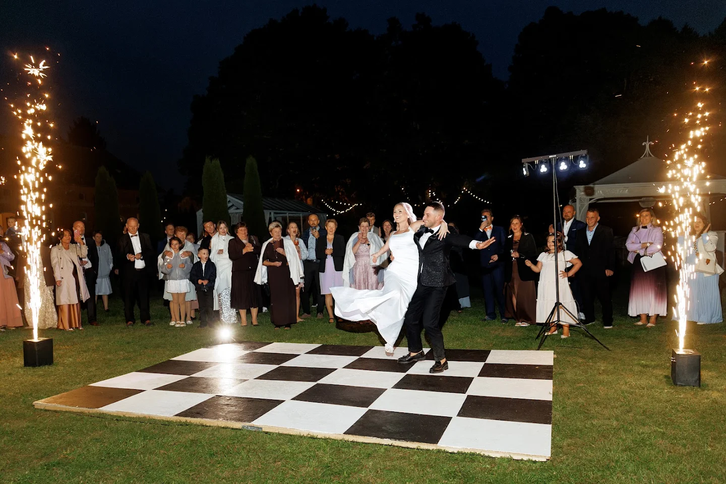 Bride and groom dancing on a black and white checkered dance floor outdoors at night with guests watching and sparklers on either side.
