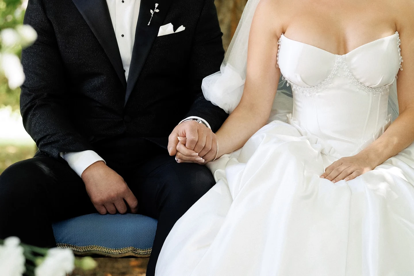 Bride and groom sitting side by side holding hands, bride wearing a white strapless wedding dress and groom in a black tuxedo.