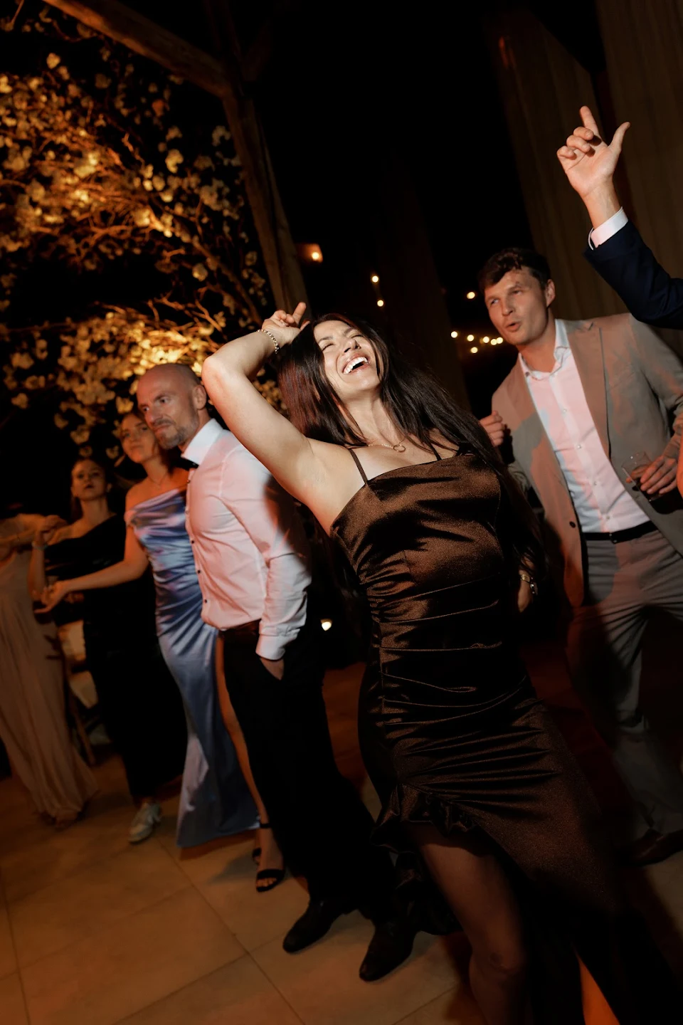 A group of people dressed formally dancing and enjoying themselves at a nighttime event with lights and a floral decoration in the background.