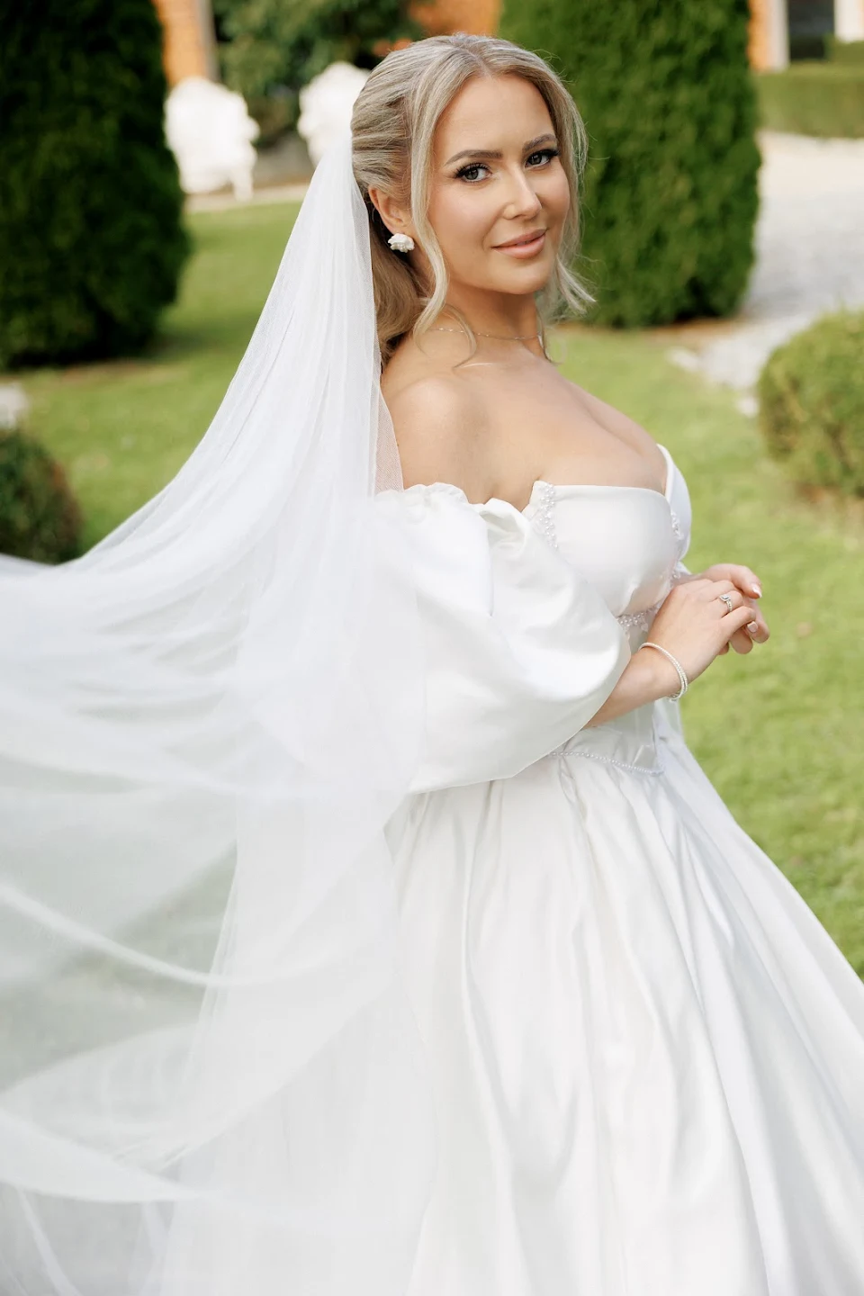 Smiling bride in an off-shoulder white wedding dress with a flowing veil outdoors.
