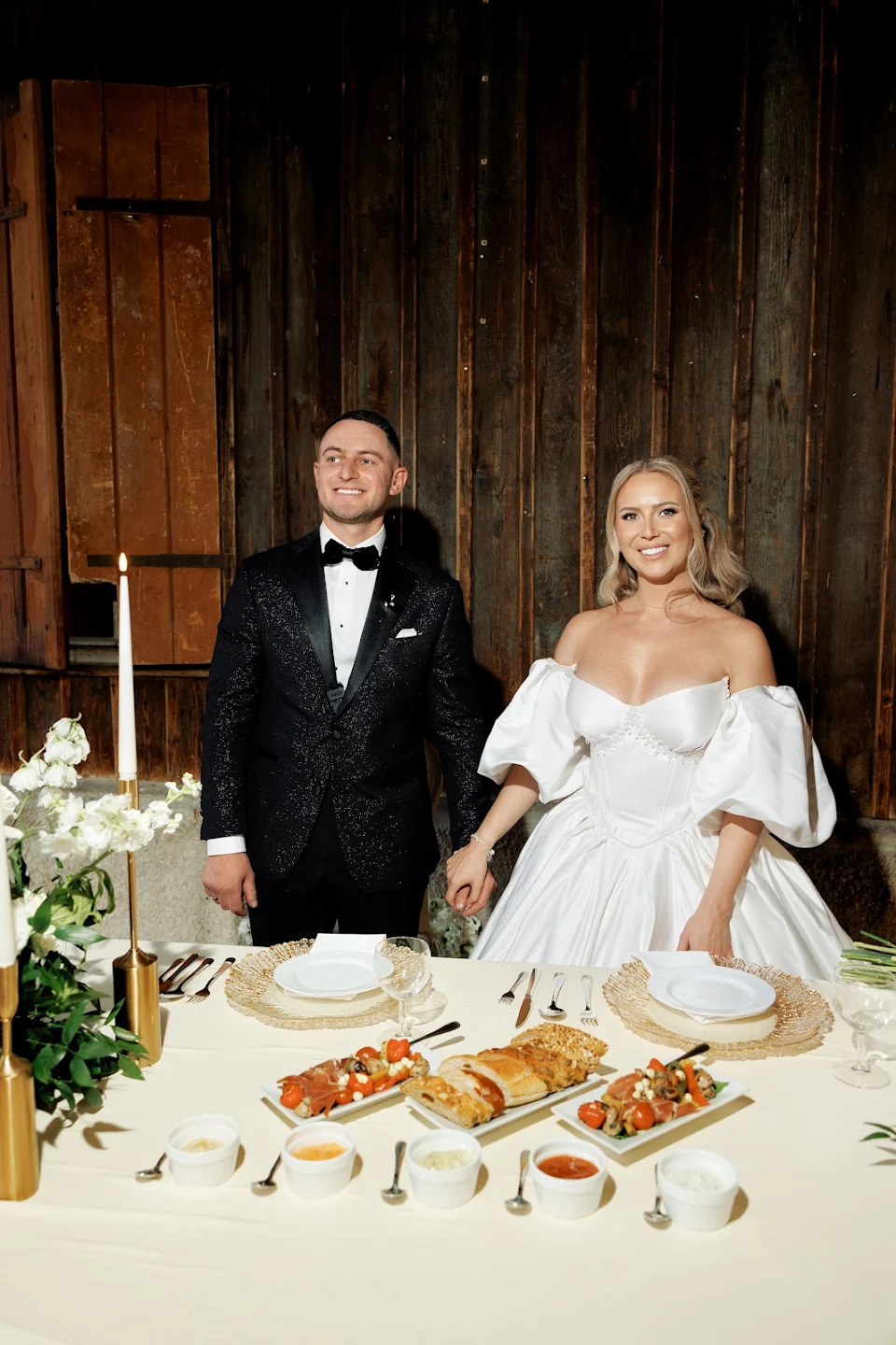 Smiling bride in white off-shoulder wedding gown and groom in black tuxedo holding hands behind a table set with bread, salads, and sauces.