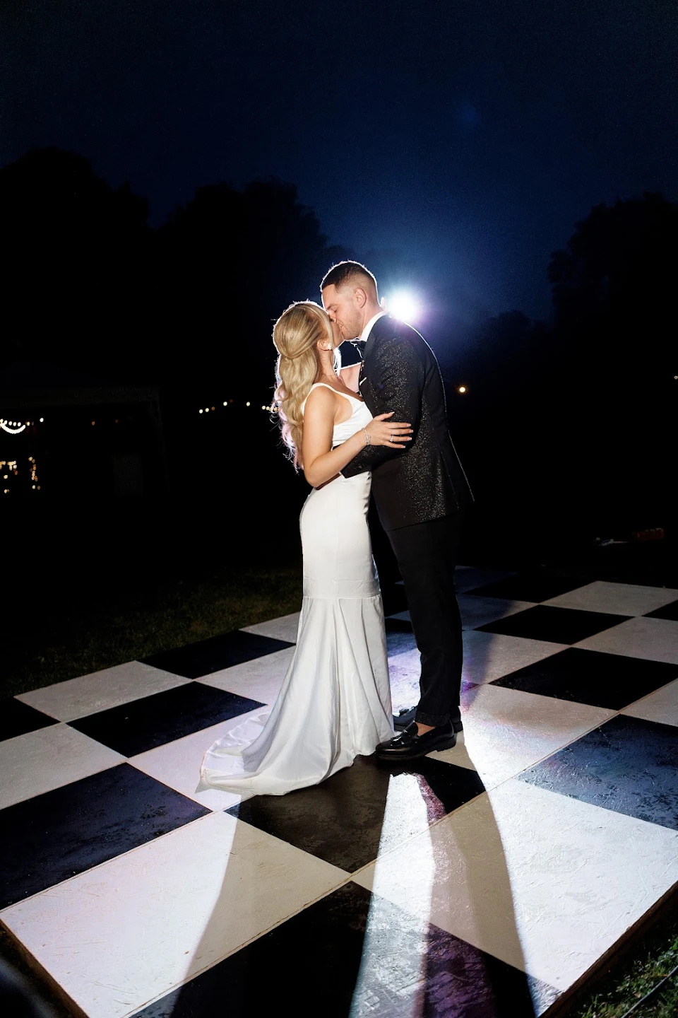 Bride and groom kissing on a black and white checkered dance floor at night with bright backlight.