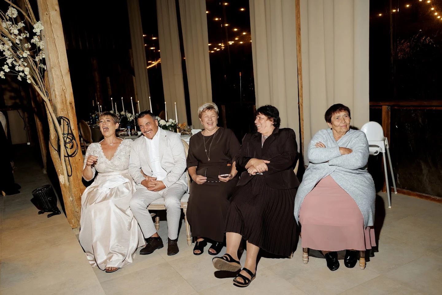 Five people sitting on chairs in a row indoors, laughing and smiling at an event at night.