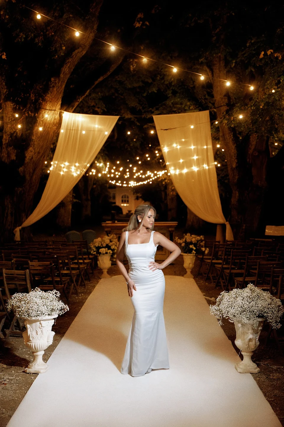 Woman in a white fitted gown posing on a white aisle runner under string lights and sheer drapes in an outdoor nighttime wedding setting.
