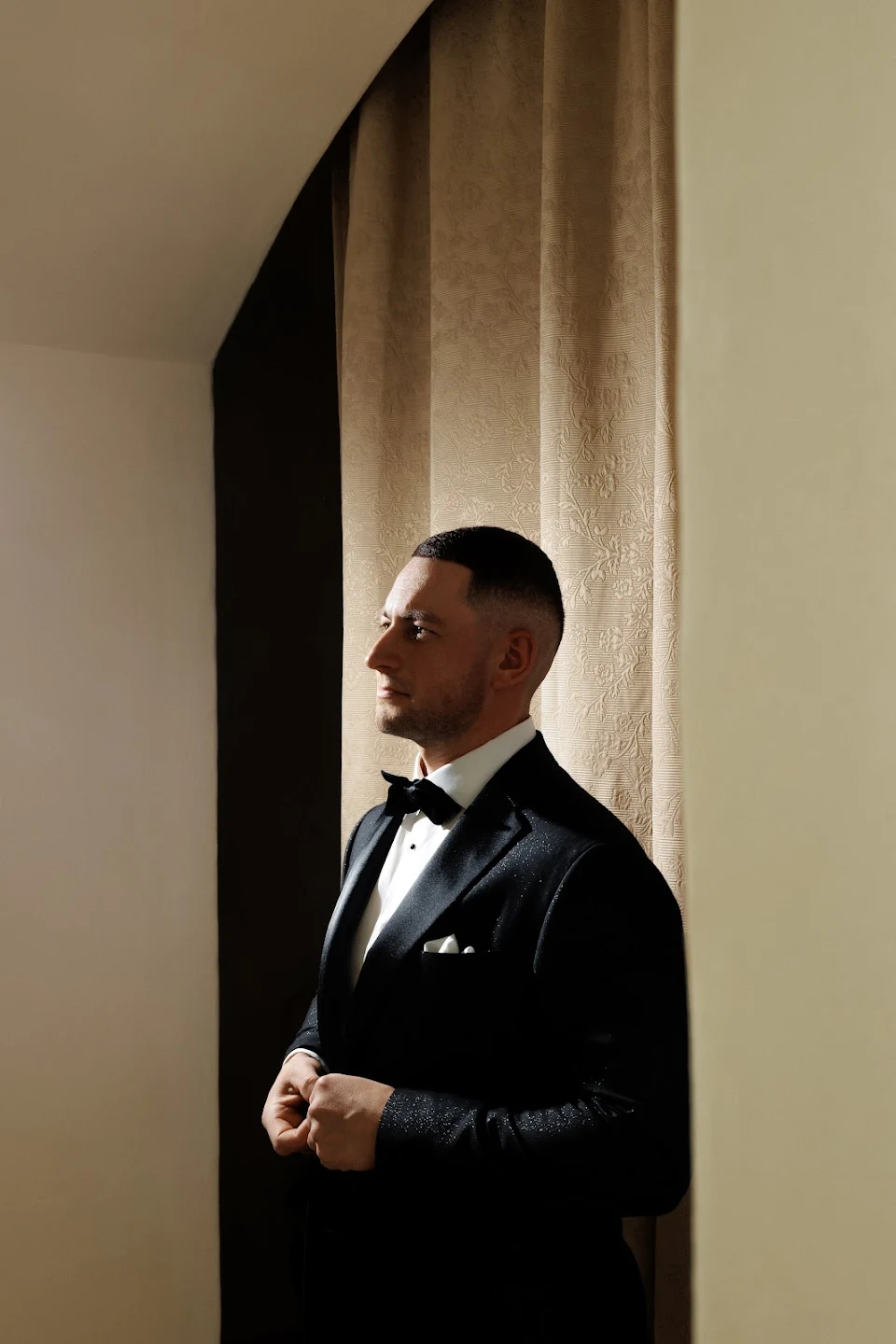 Man in a black tuxedo with a bow tie standing sideways near a beige curtain.