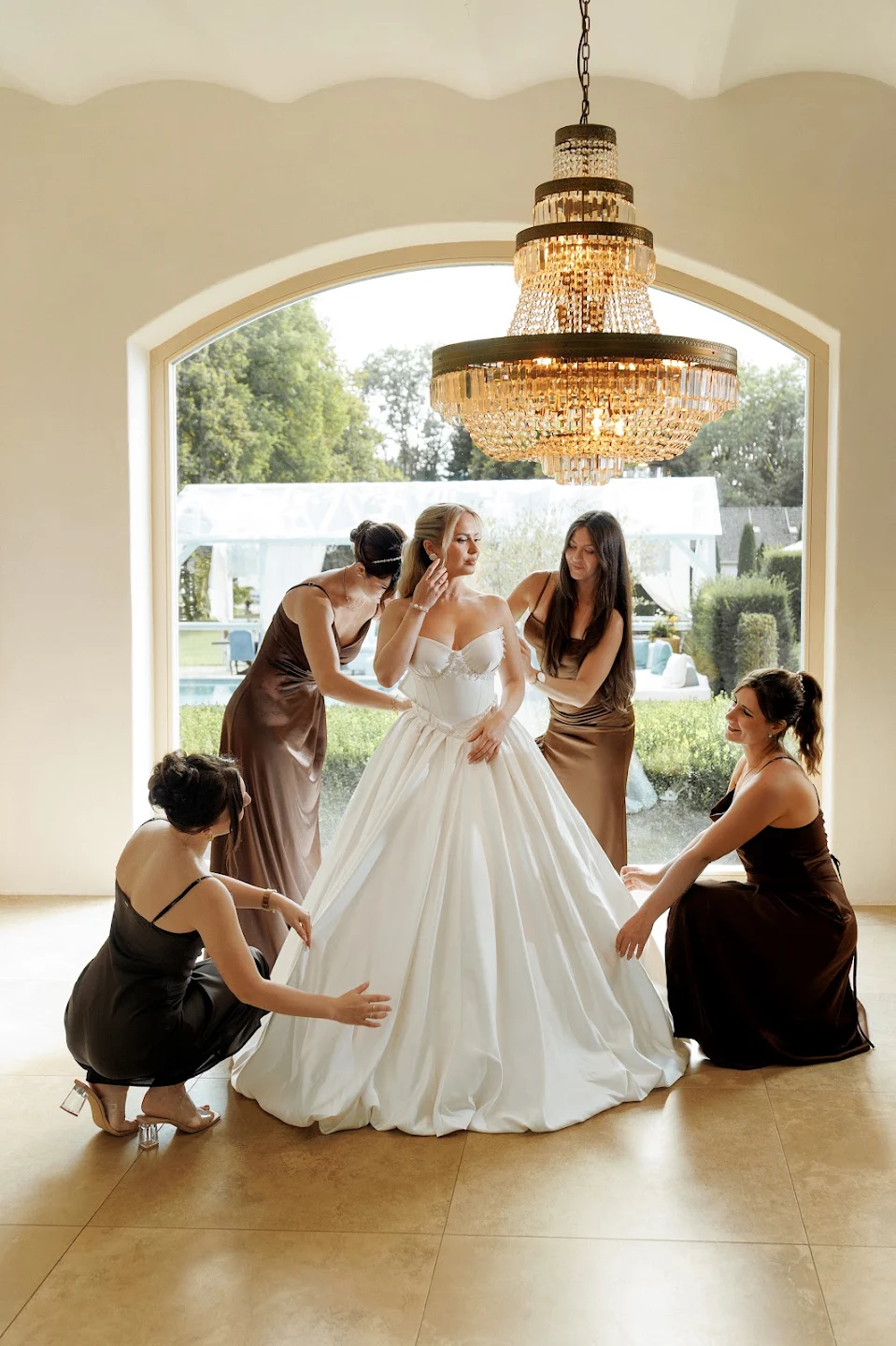 Bride in a white wedding gown being helped by four bridesmaids wearing brown dresses near a large arched window with a chandelier overhead.