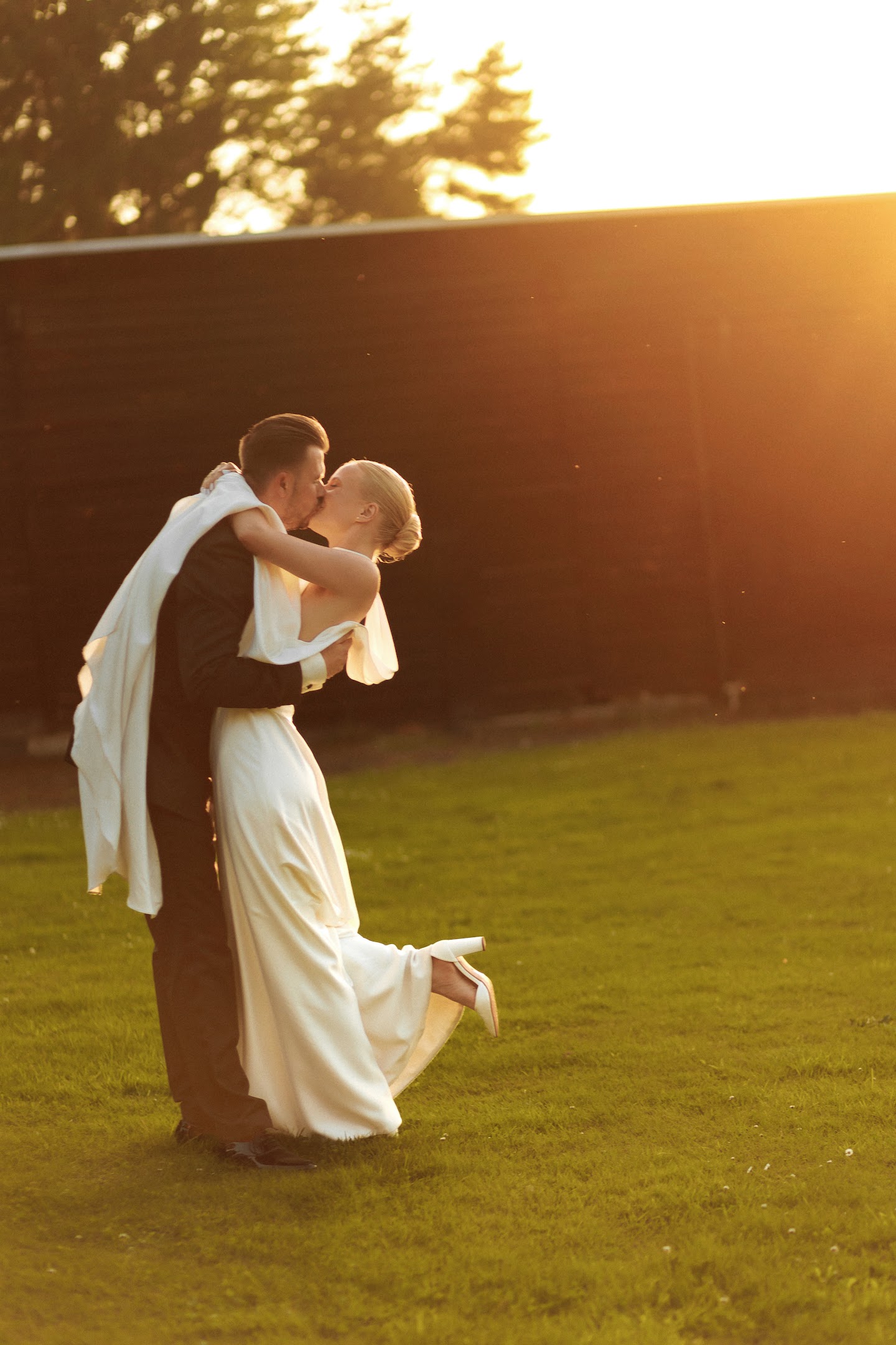 Bride in a white dress and groom in a dark suit kissing outdoors on green grass at sunset.