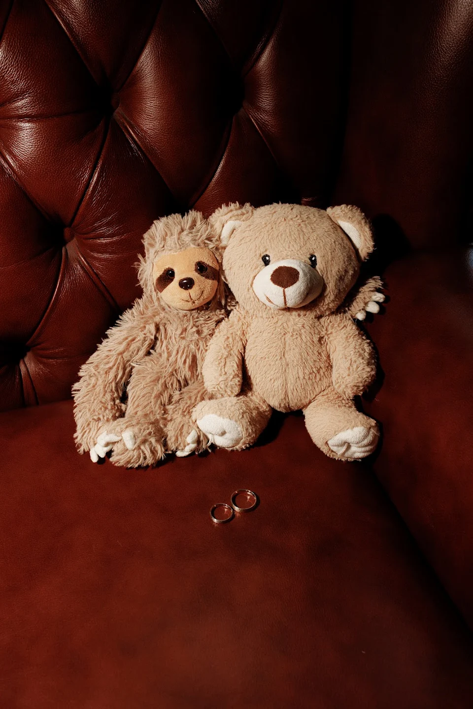 Two plush toys, a sloth and a teddy bear, sitting closely together on a brown leather couch with two gold rings placed in front of them.