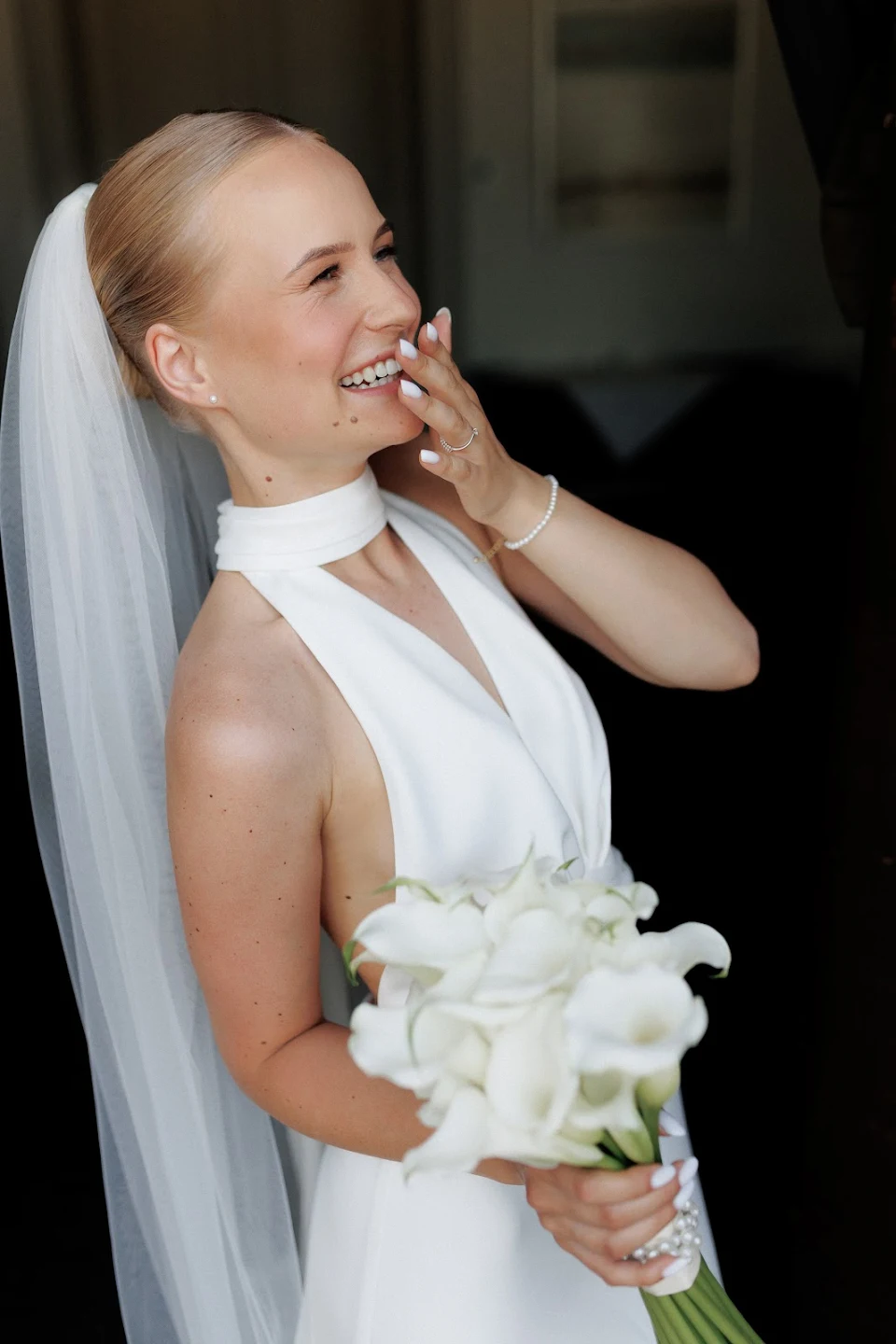 Smiling bride in a white halter dress and veil holding a bouquet of white flowers.
