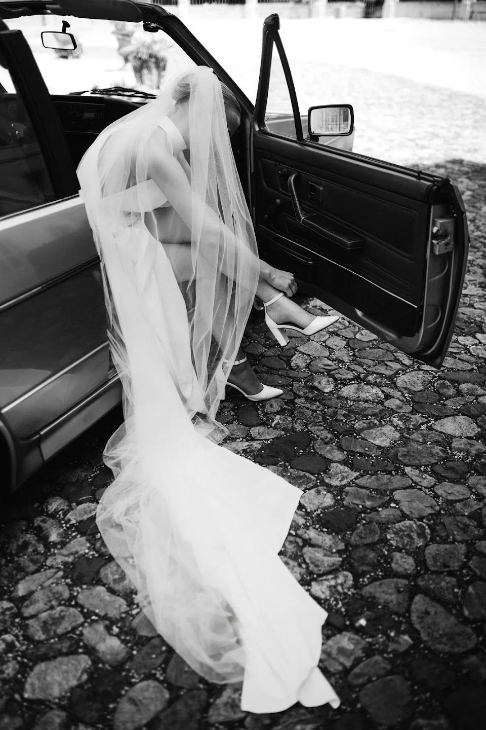 Black and white photo of a bride sitting in a car while fastening the strap of her high-heeled shoe, wearing a long veil and wedding dress.