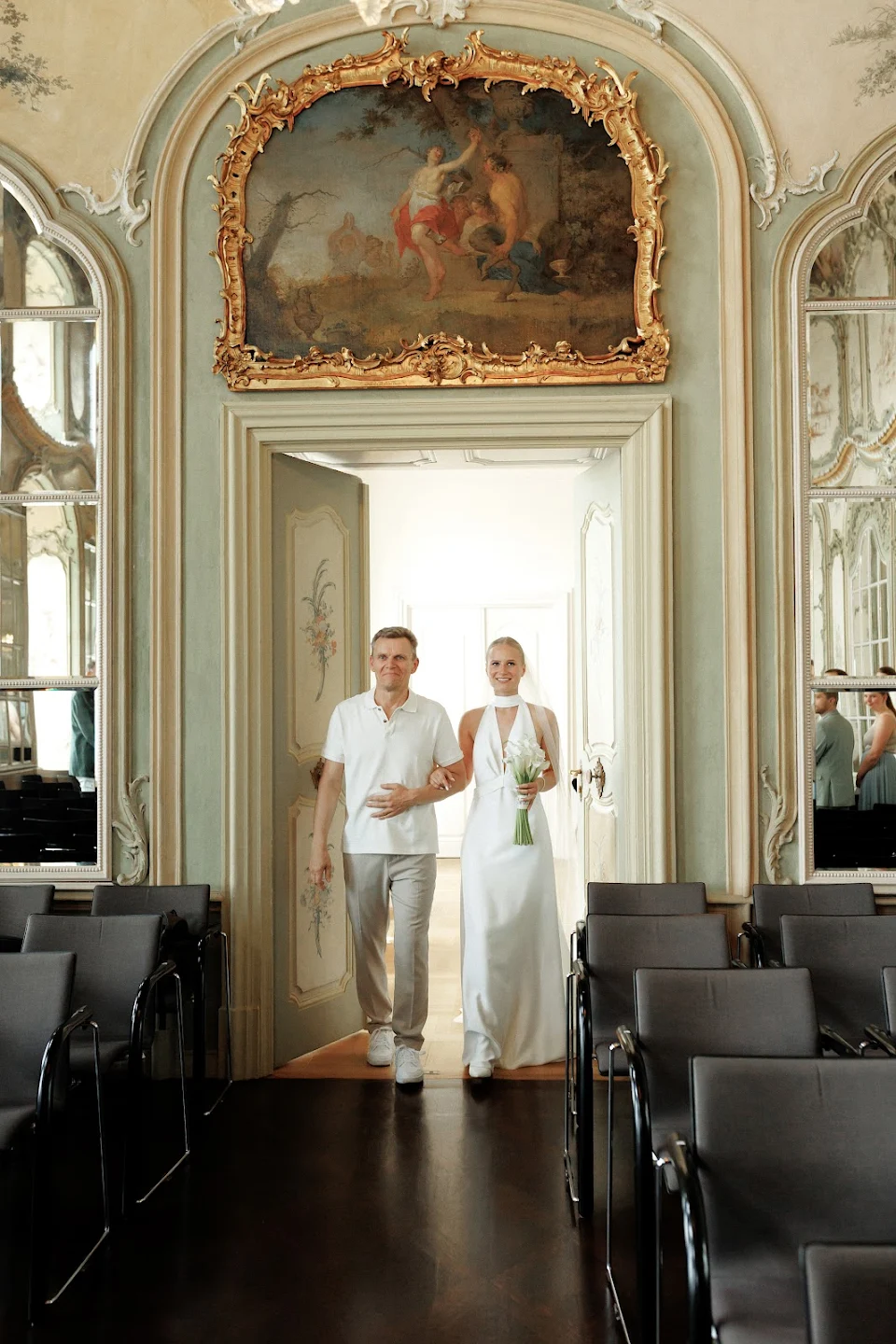Bride in white gown holding flowers walking arm-in-arm with a man in casual attire through ornate double doors in an elegant room.