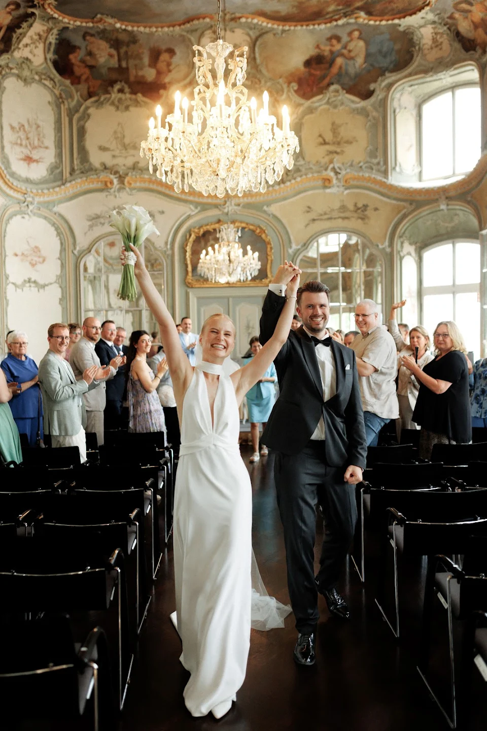 Bride and groom smiling and holding hands while walking down the aisle in an ornate room with a chandelier as guests applaud.