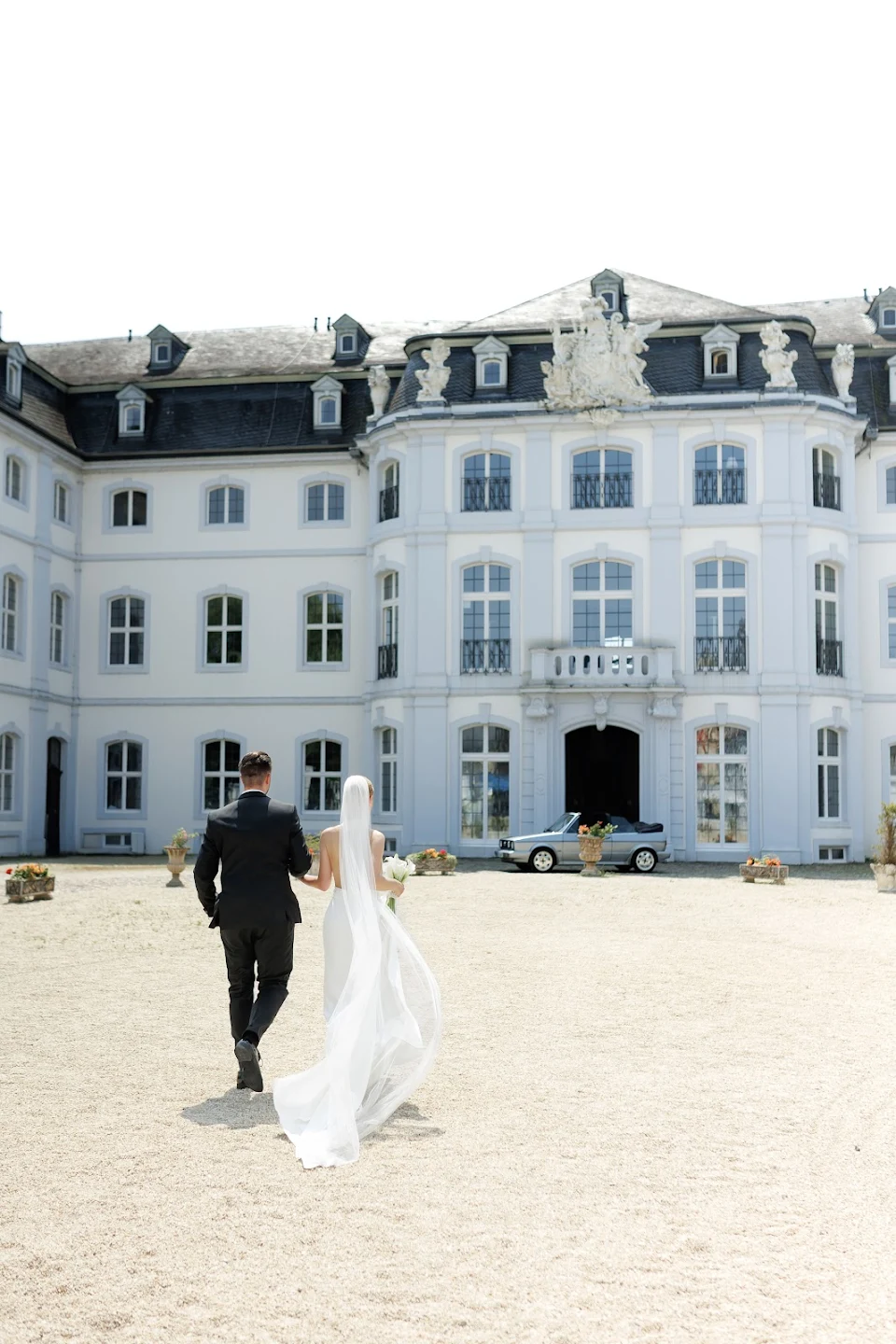 Bride and groom walking on gravel towards a large white mansion with many windows on a sunny day.