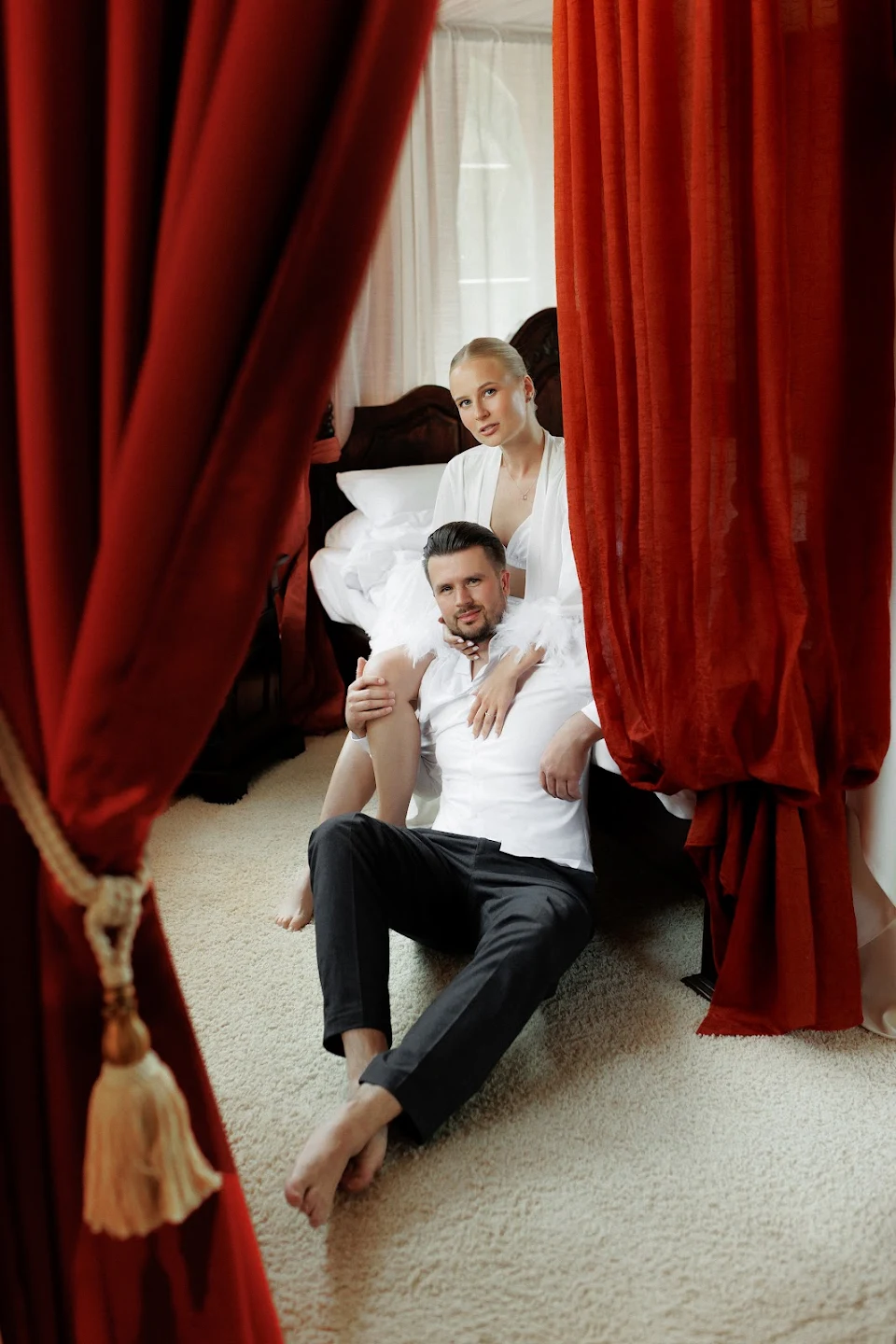 Couple dressed in white sitting on a carpeted floor, framed by red curtains, with a wooden bed and white bedding in the background.