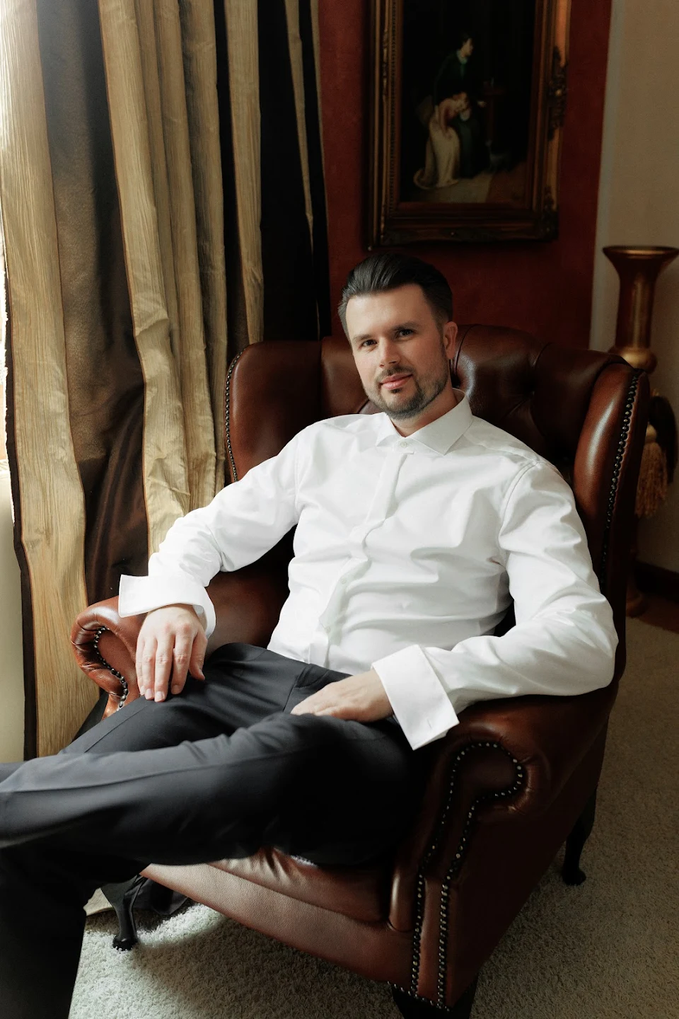 Man in white dress shirt and dark pants sitting relaxed in a brown leather armchair by a window with beige curtains.