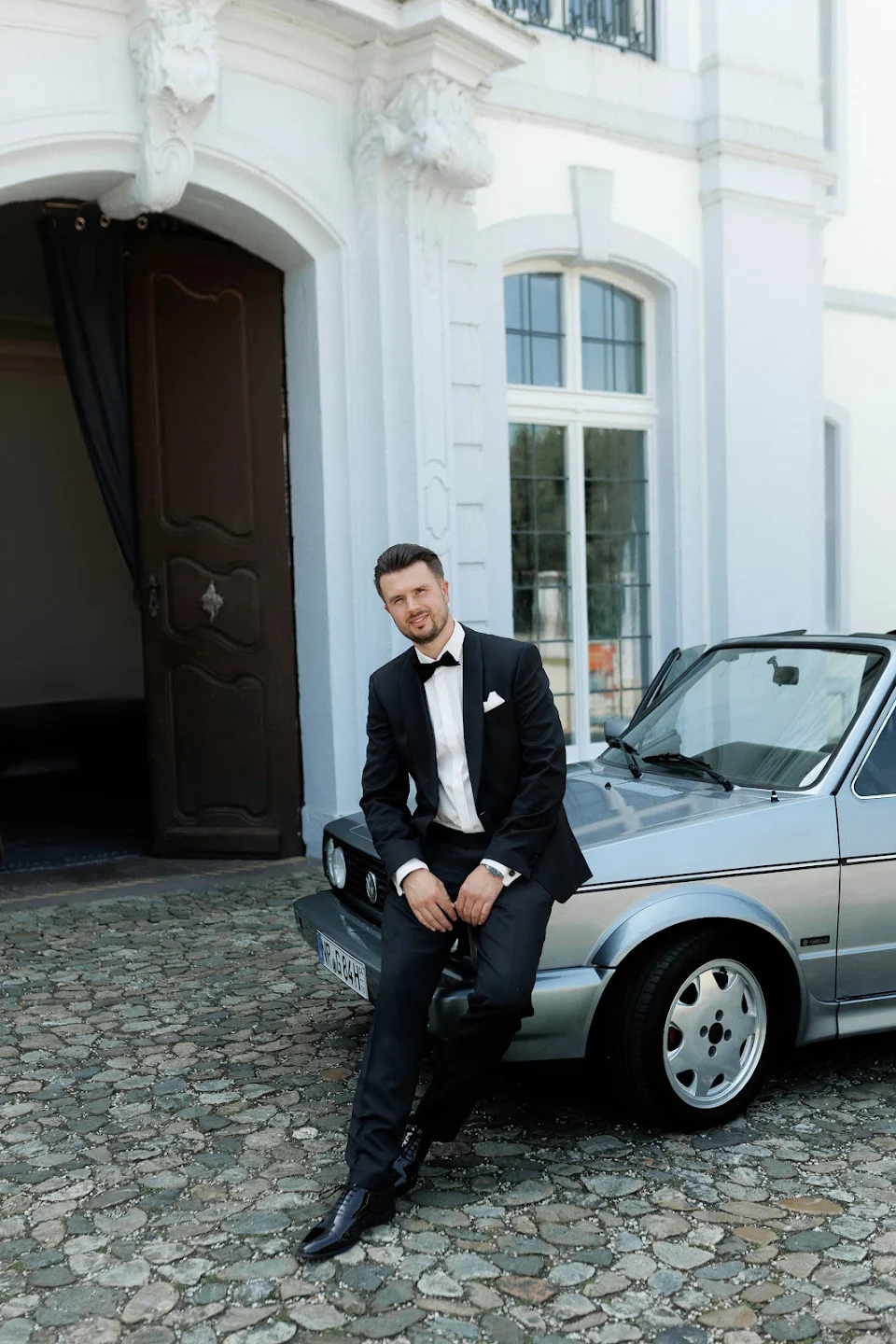 Man in black tuxedo sitting on the front bumper of a silver vintage convertible car outside a white ornate building.