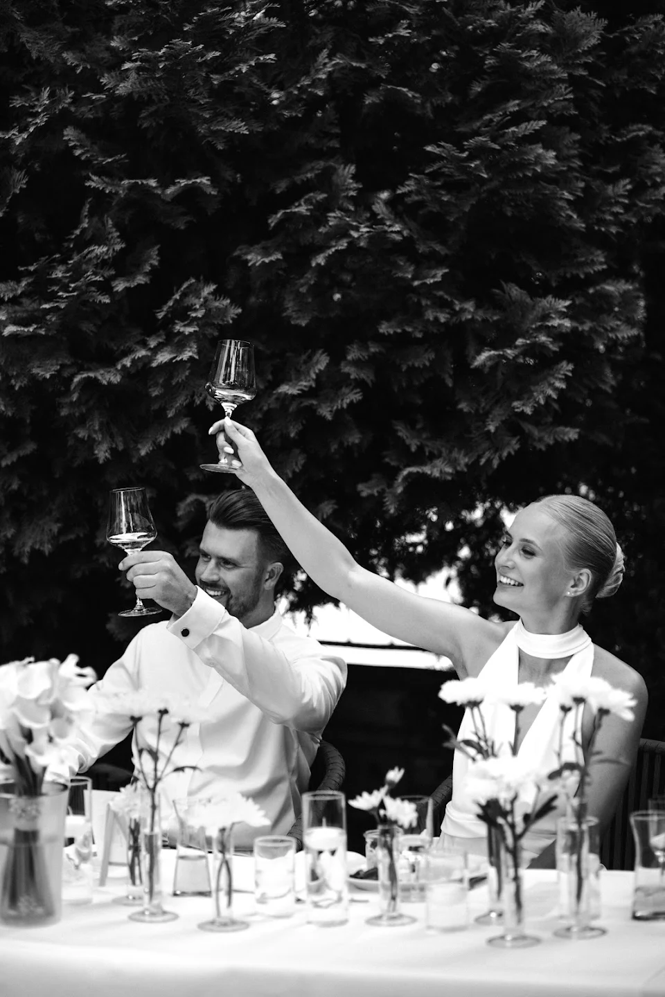 A man and woman dressed in white raising wine glasses in a celebratory toast at a table decorated with flowers.