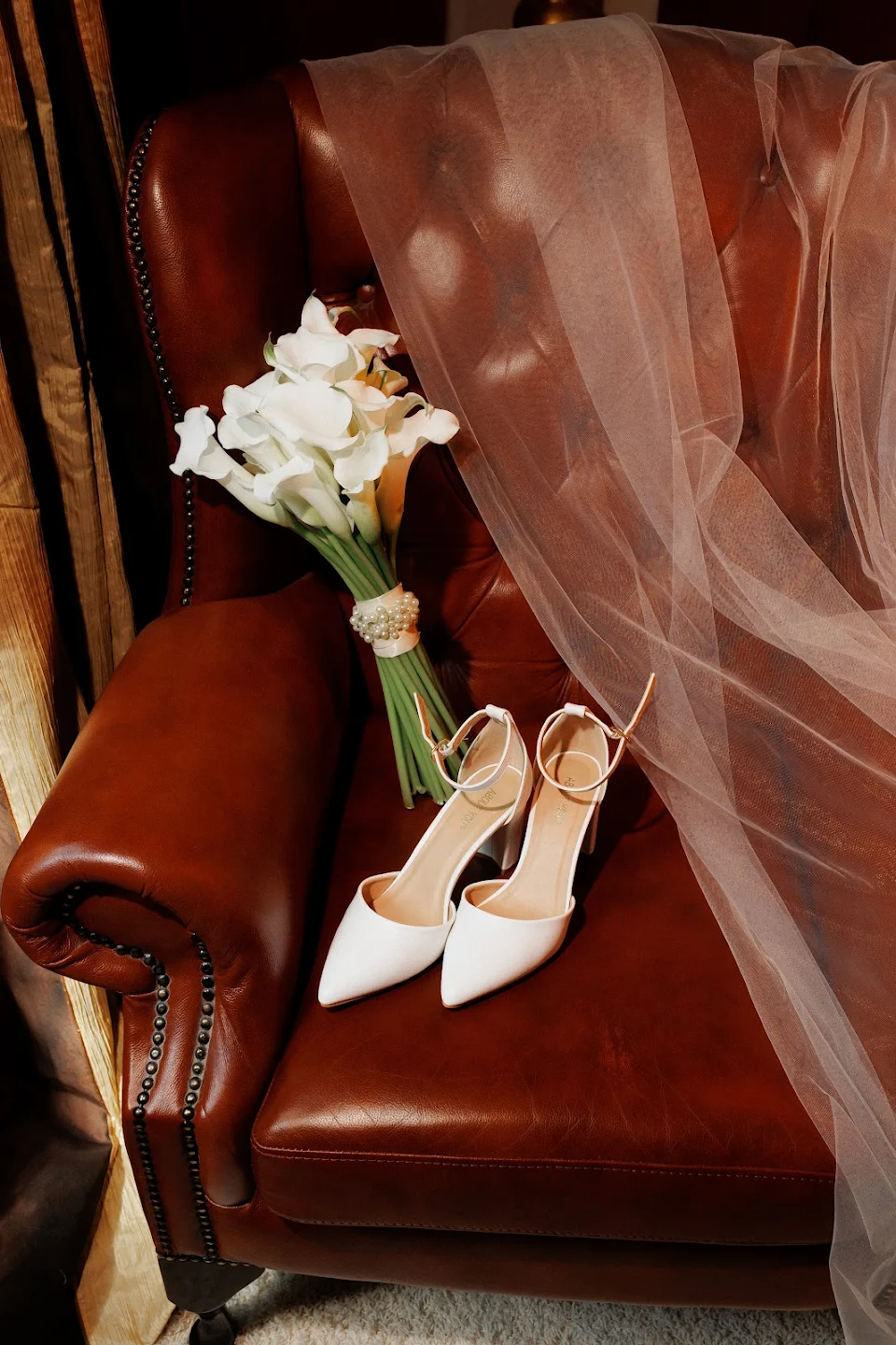 White bridal heels, bouquet of white calla lilies, and a sheer veil draped on a brown leather armchair.