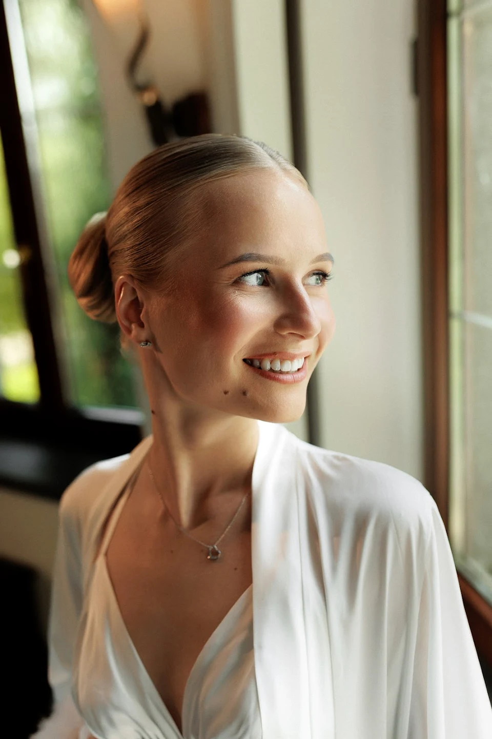 Smiling woman with blonde hair in a bun wearing a white blouse and silver necklace standing by a window.