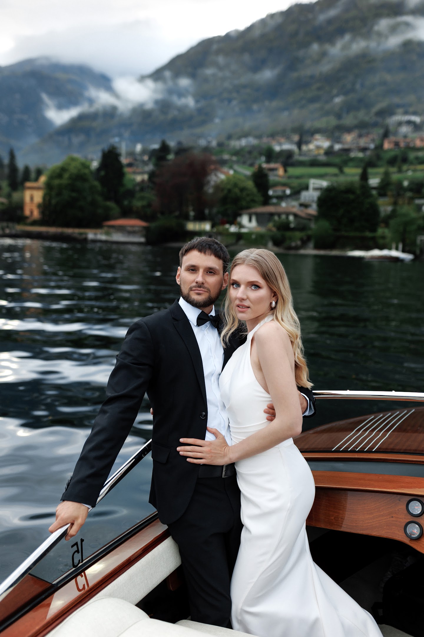 A formally dressed couple embracing on a wooden boat with a lake and mountains in the background.