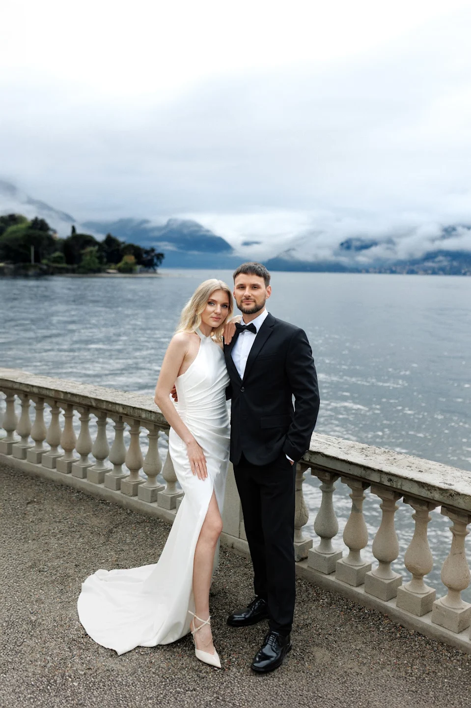 A couple dressed formally with the woman in a white gown and the man in a black suit standing on a terrace by a lake with mountains and cloudy sky in the background.
