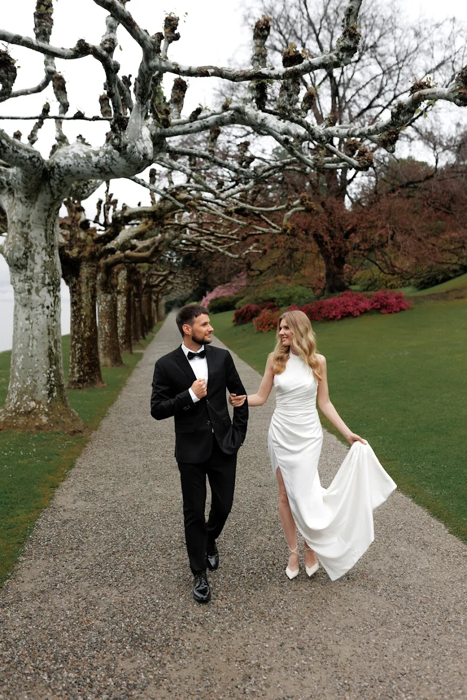Couple dressed in formal wedding attire walking arm in arm down a tree-lined gravel path in a park.