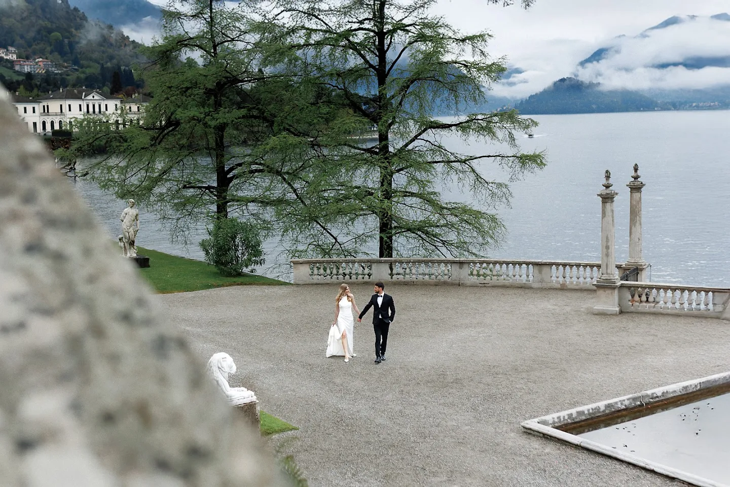 A bride in a white dress and a groom in a black tuxedo walking hand in hand on a riverside terrace with trees, statues, and mountains in the background.
