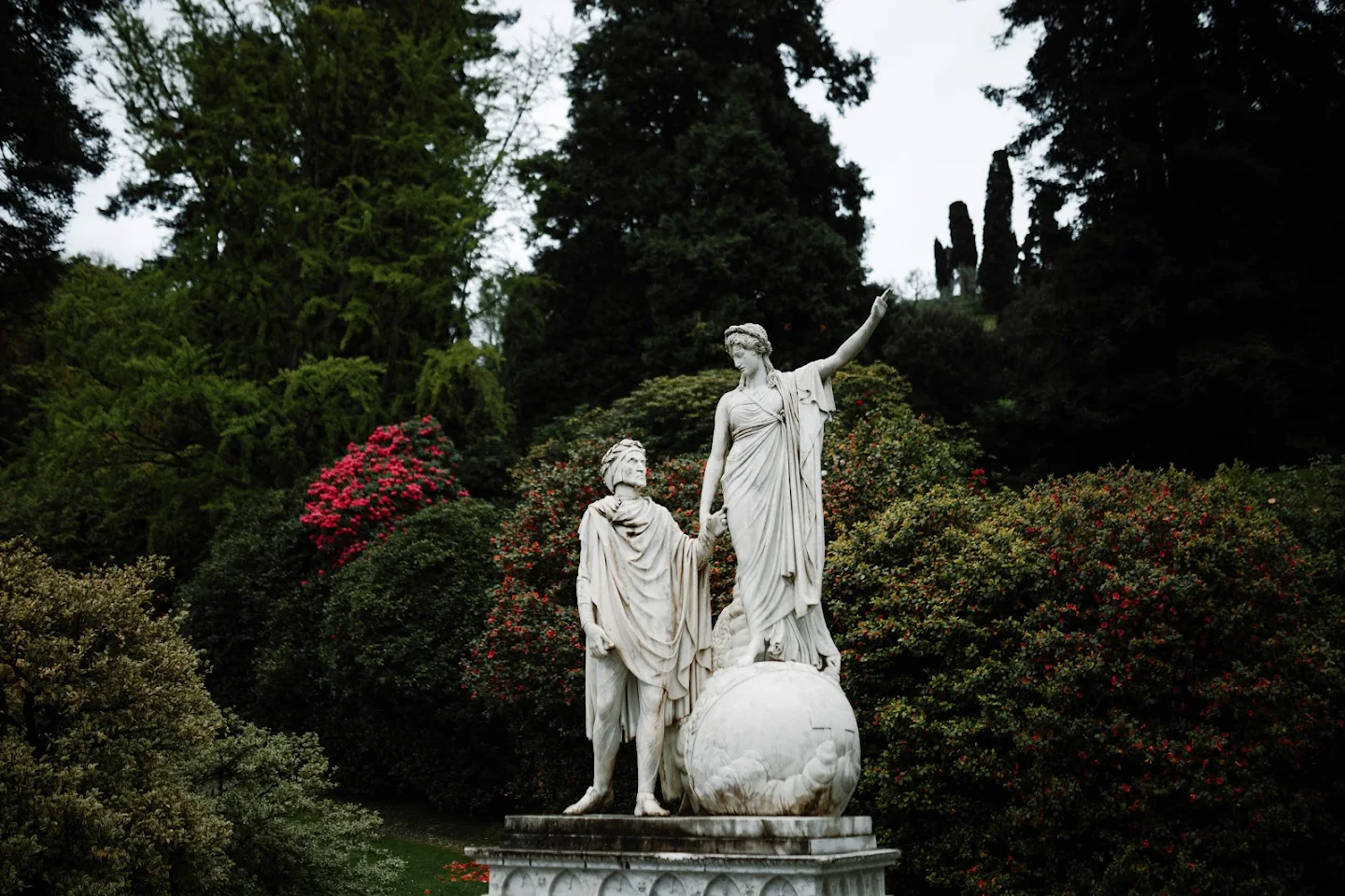 Marble statue of a man and a woman holding hands, with the woman standing on a globe, surrounded by lush greenery and blooming shrubs.