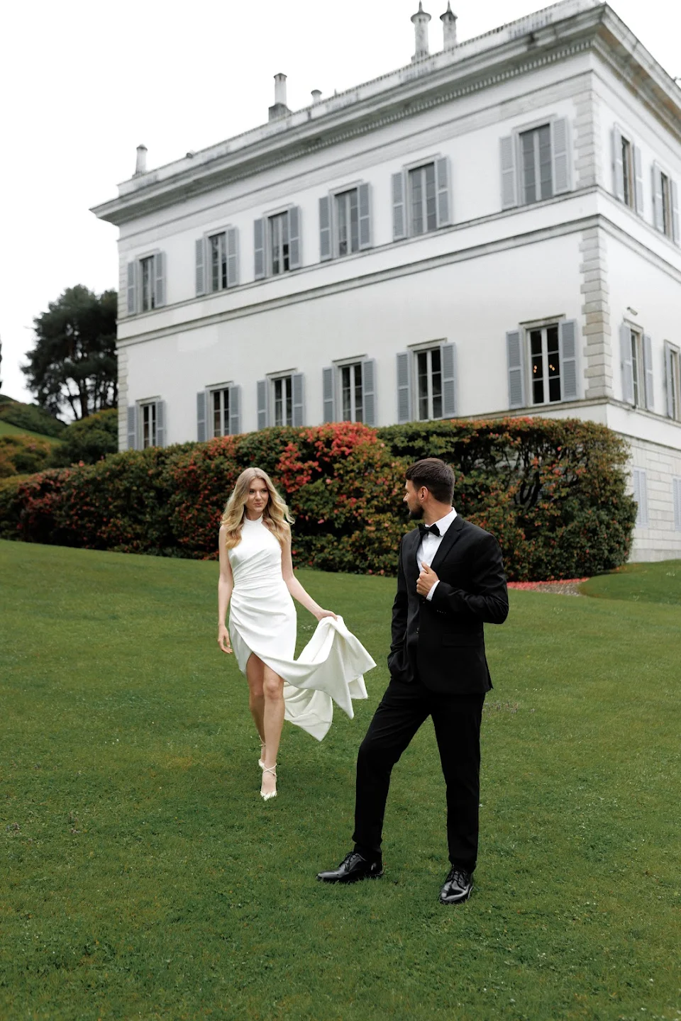 A woman in a white dress walking on green lawn towards a man in a black tuxedo standing near a large white building with grey shutters.