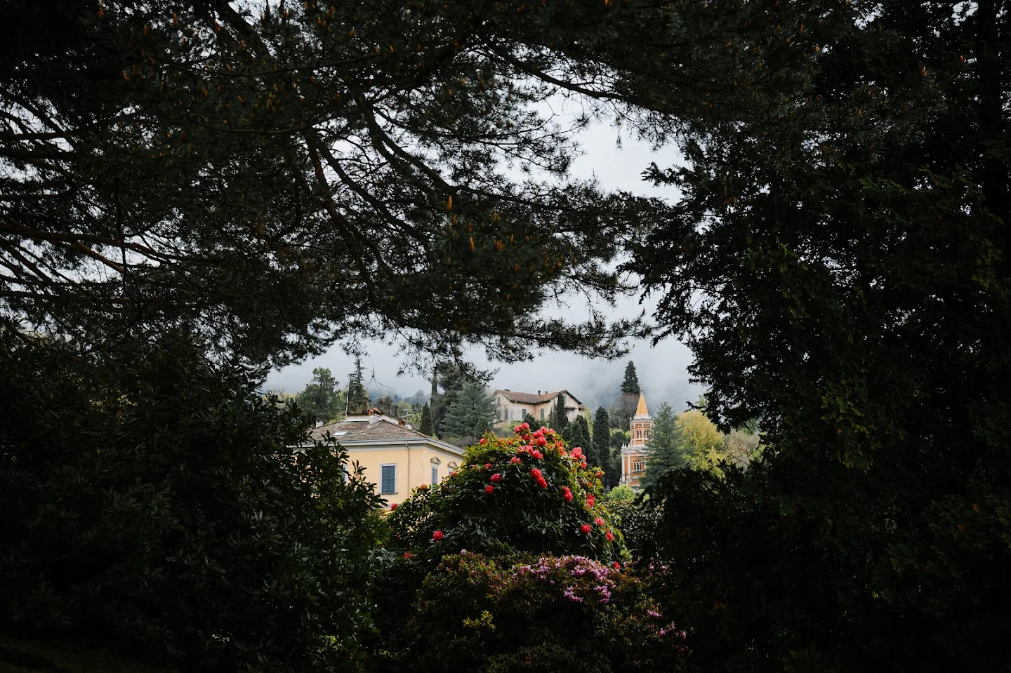 View of a yellow house and tall tower partially obscured by dense trees and vibrant flowering bushes under a cloudy sky.