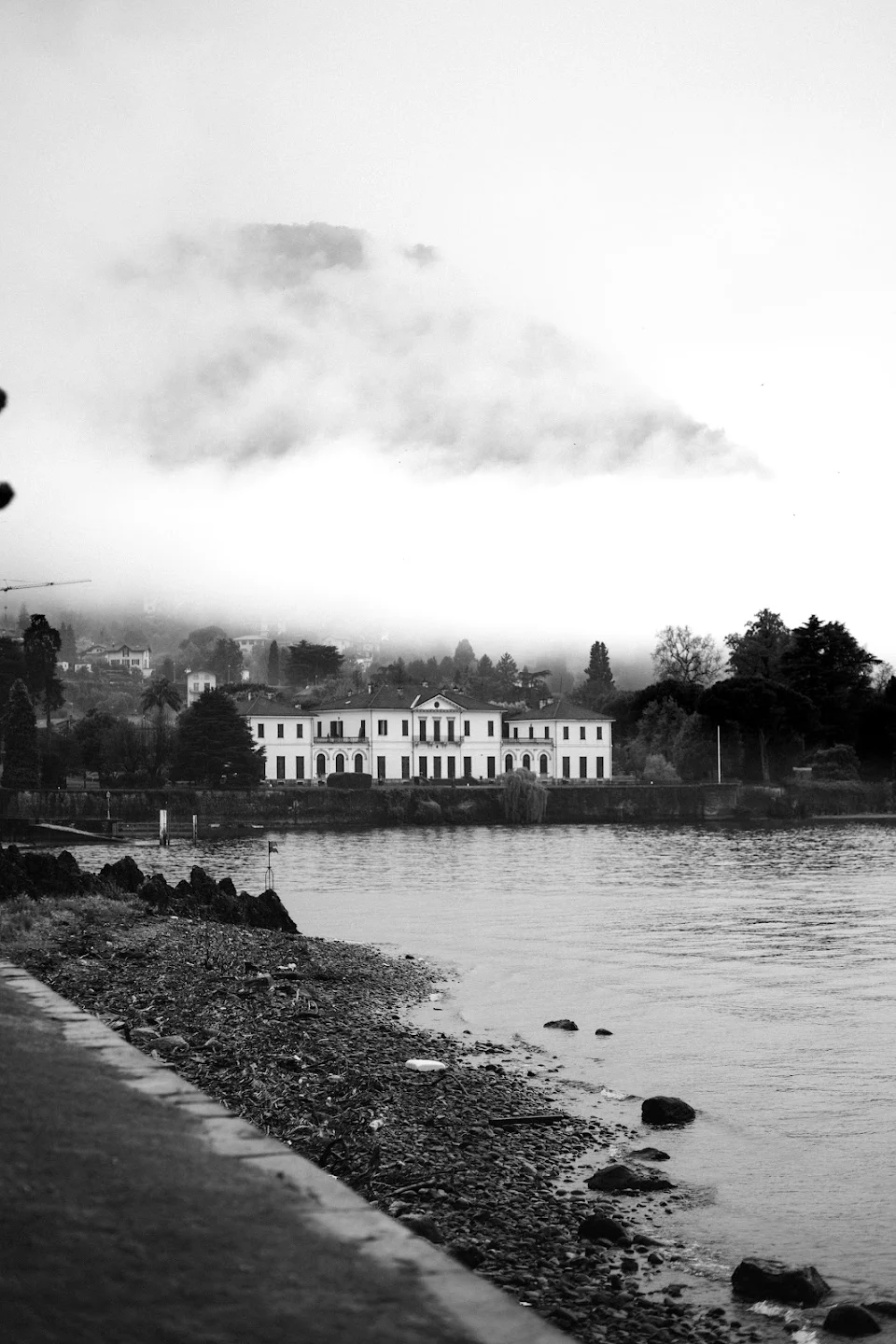 Black and white photo of a large white building by a lakeshore with mist-covered hills in the background.