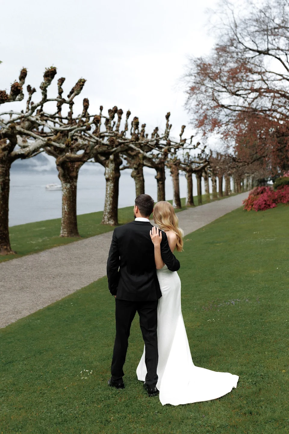 Bride and groom standing arm in arm on a grassy path lined with pruned trees, looking out towards a body of water.
