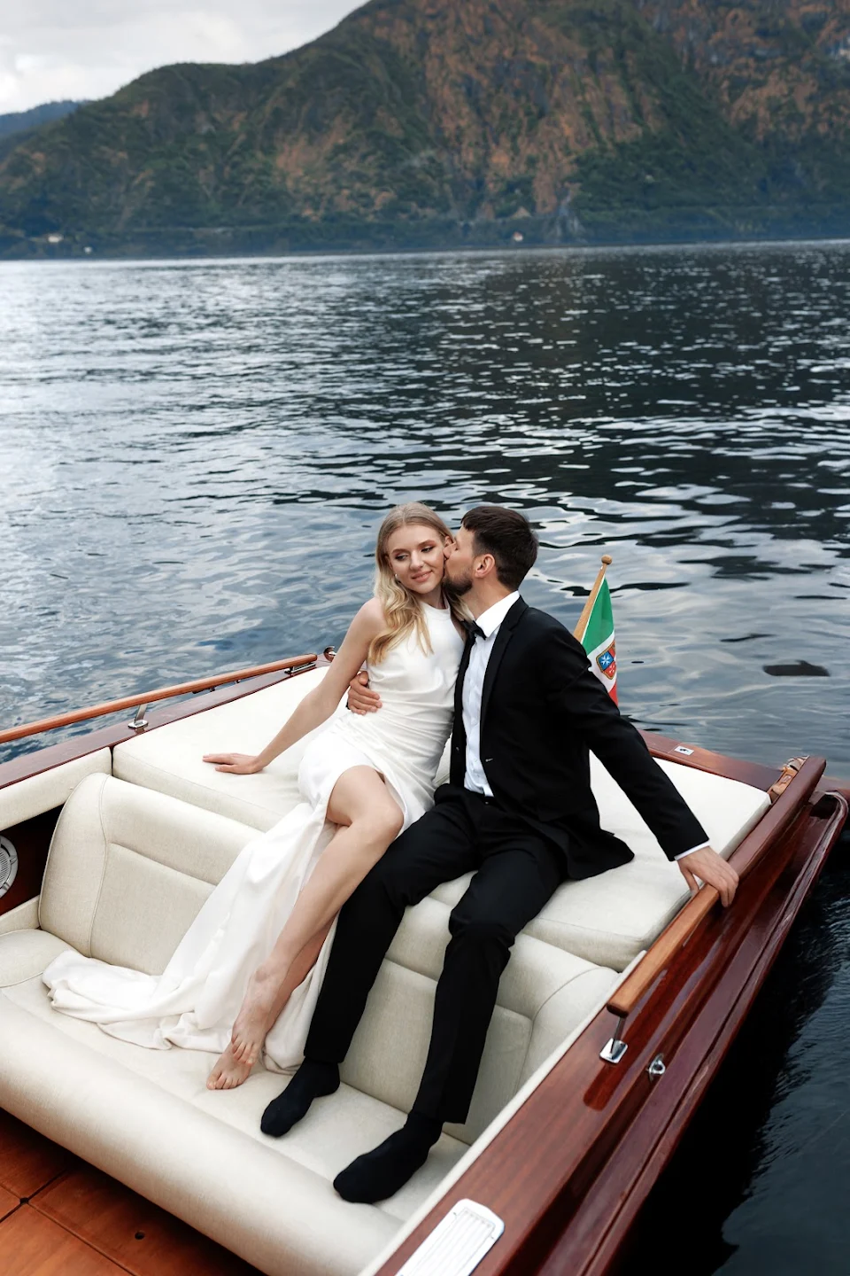 Couple dressed elegantly with the man kissing the woman on the cheek while sitting on a boat on a lake with mountains in the background.