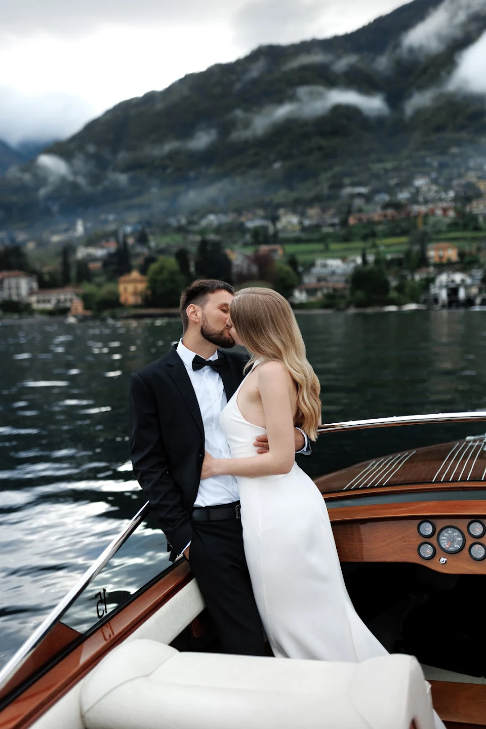 Couple dressed in wedding attire kissing on a boat with a mountainous lakeside village in the background.