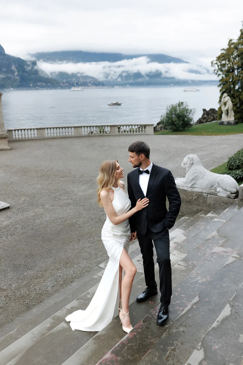Couple in elegant wedding attire standing on outdoor stone steps with lake and mountains in the background.