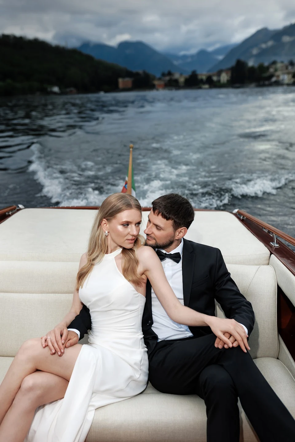 A couple dressed elegantly with the woman in a white dress and the man in a black suit sitting closely on a boat with water and mountains in the background.