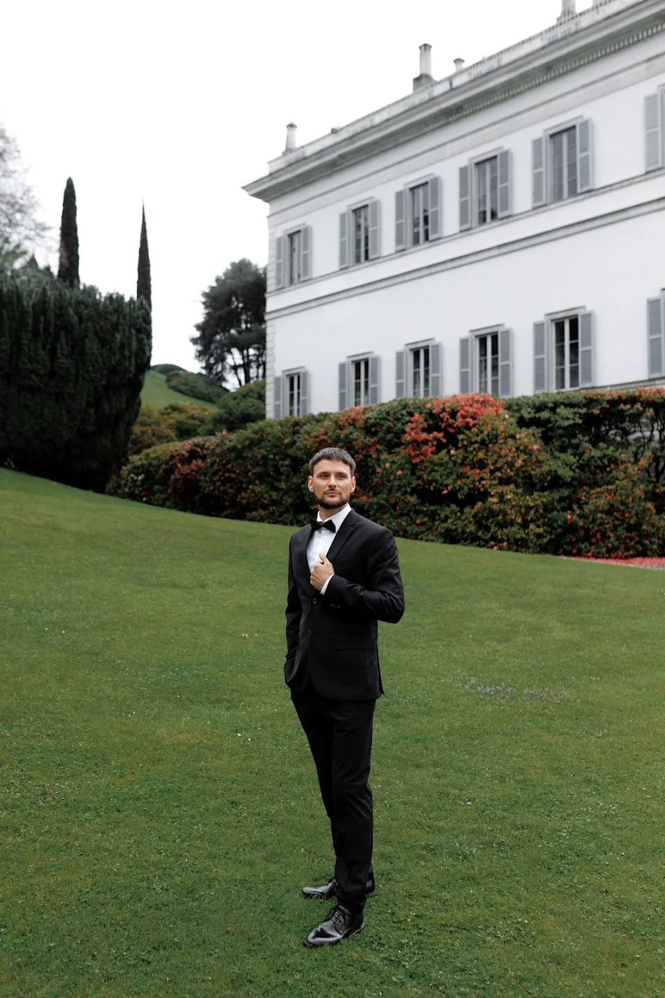 Man in a black tuxedo standing on green lawn with white building and bushes in the background.