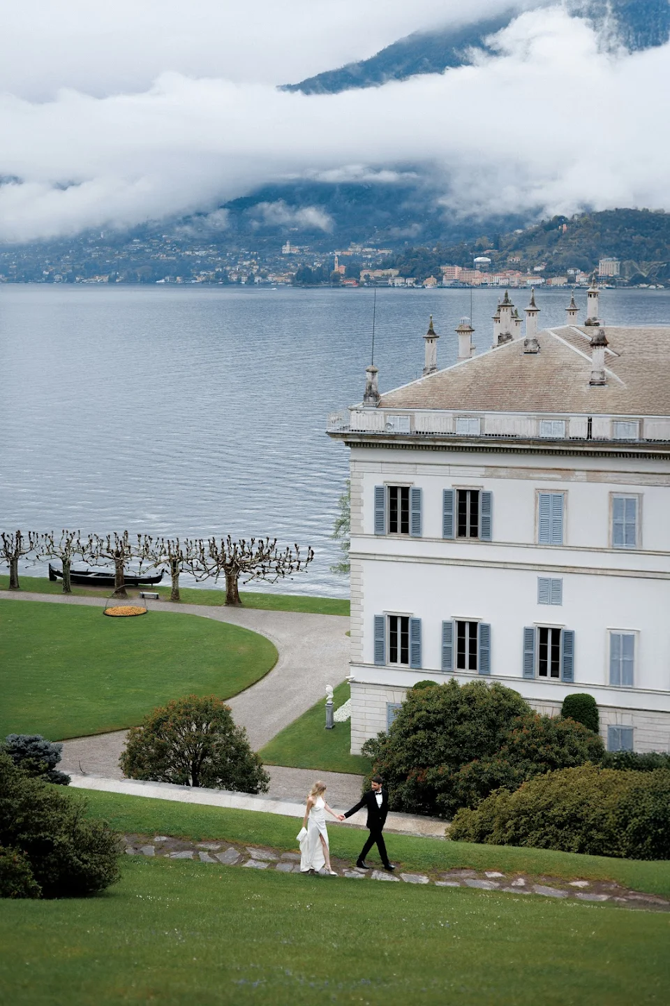 Couple in wedding attire walking hand in hand on a grassy path beside a large white building overlooking a lake with misty mountains in the background.