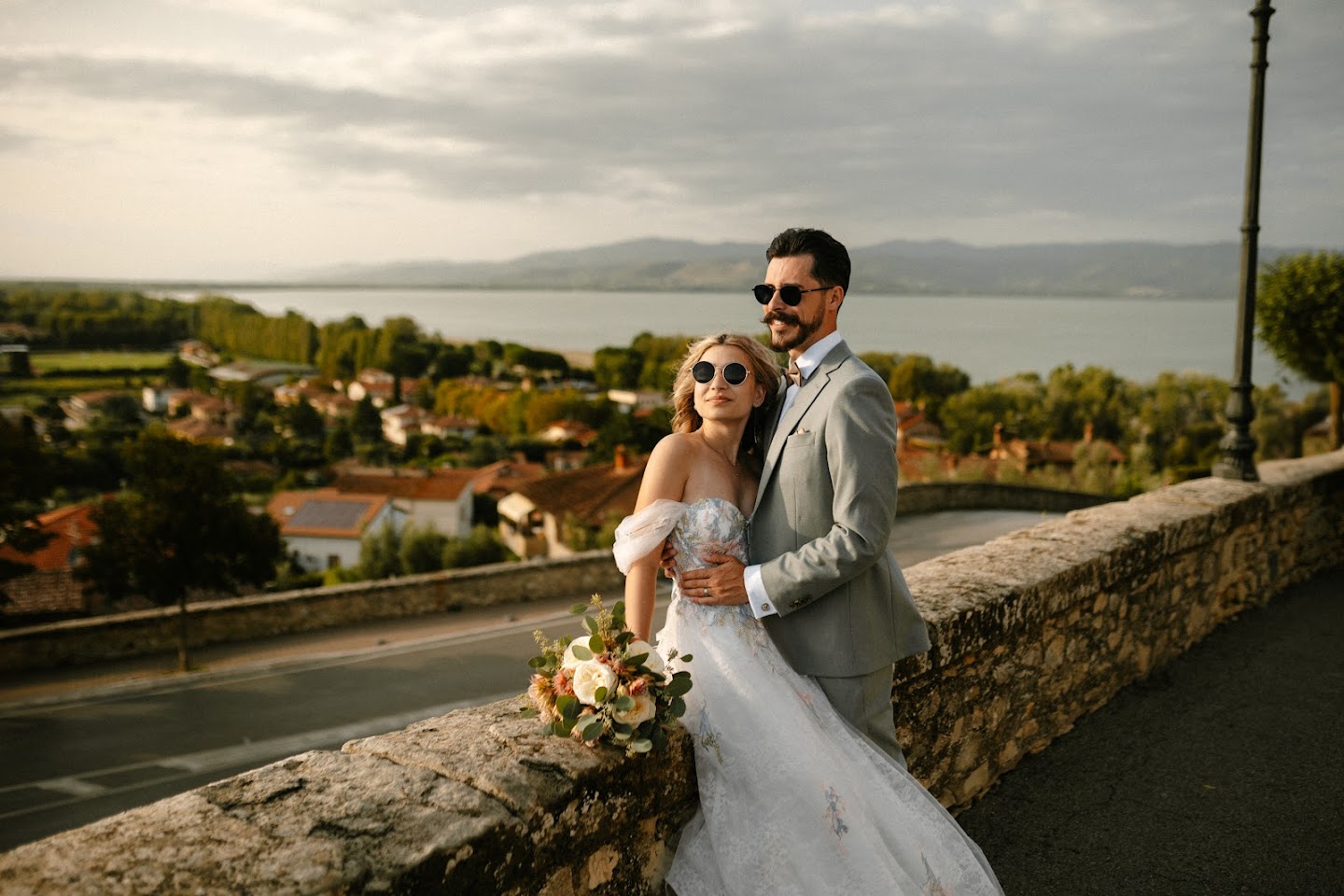 Bride and groom wearing sunglasses embracing on a stone wall overlooking a scenic town with a lake and mountains in the background.